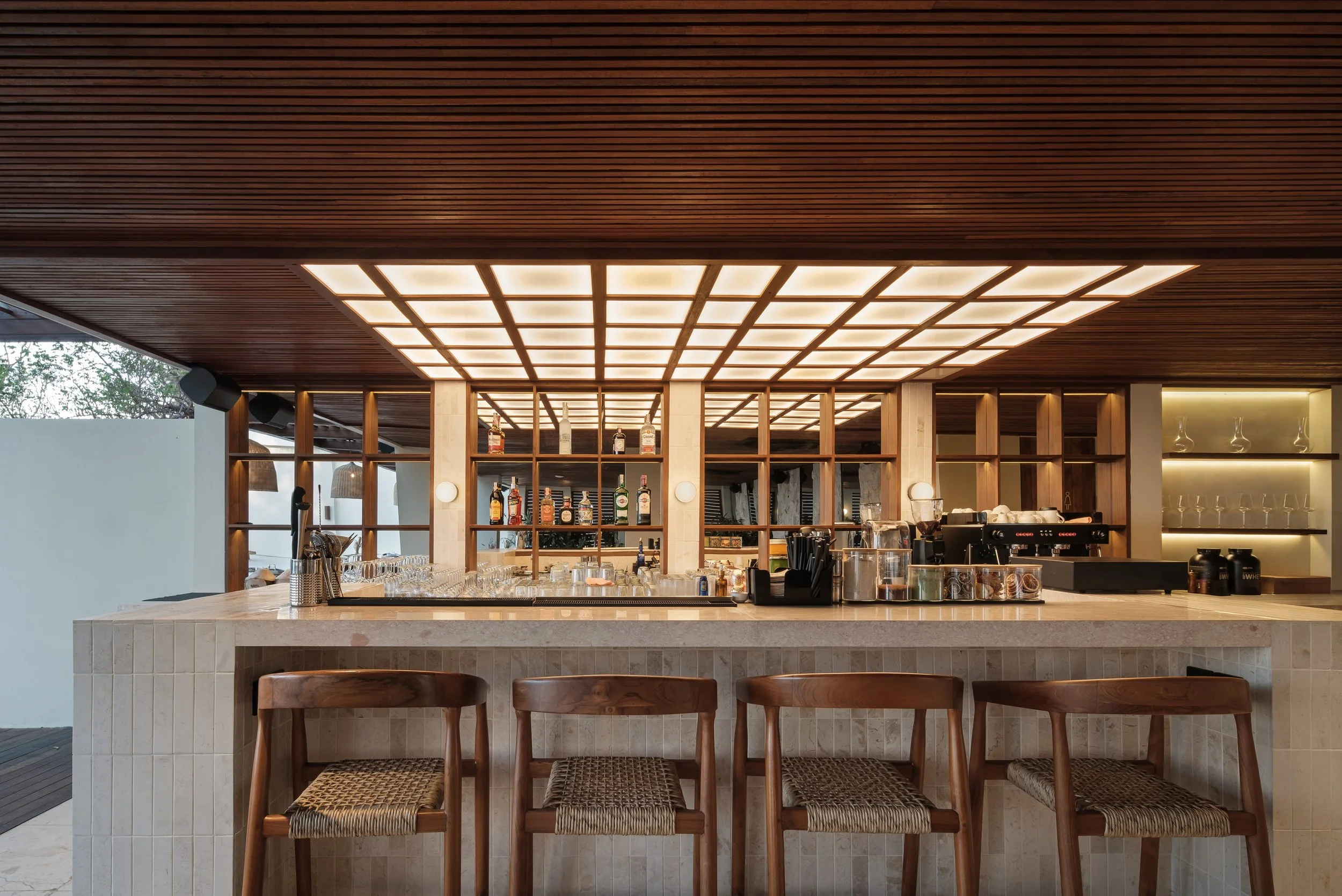 Interior view of a modern bar with a marble countertop, wooden bar stools, wooden shelving with bottles of alcohol, and a ceiling with grid-patterned lighting.
