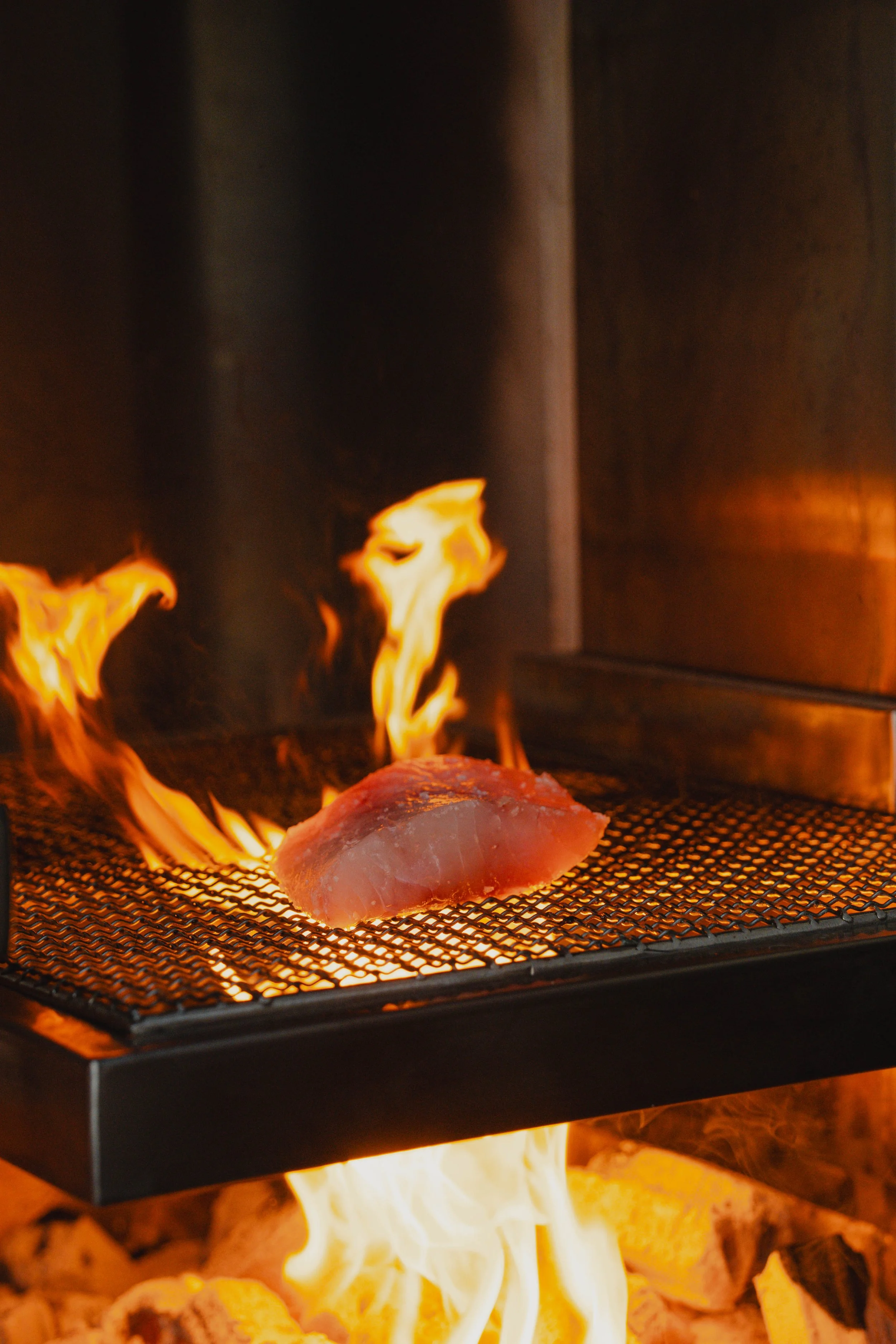 A piece of raw fish on a grill with flames and heat beneath, possibly in a restaurant or kitchen setting.