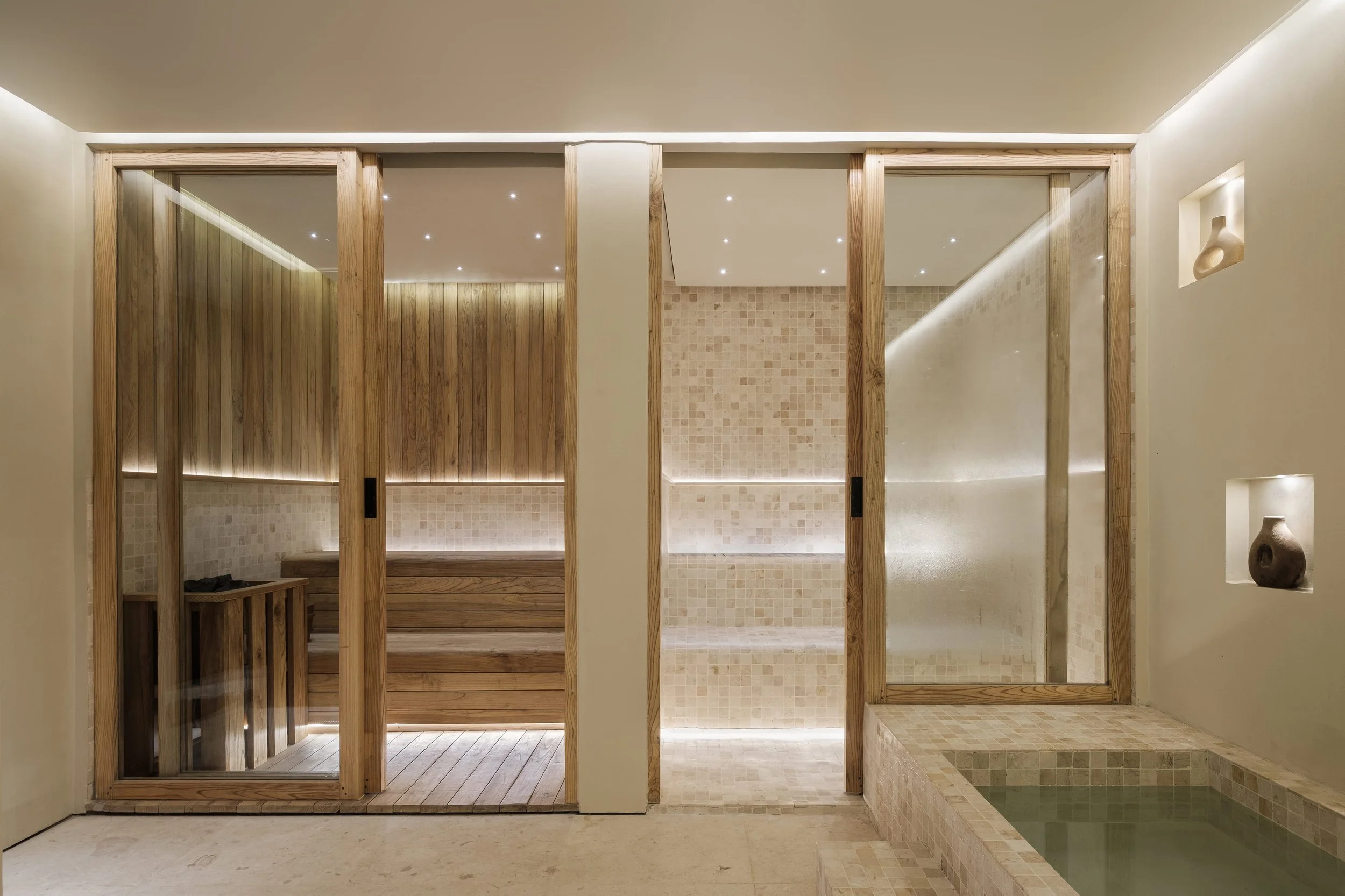 A modern spa room with wooden partition sliding doors, a tiled shower area, and a small soaking pool. The room features minimalist decor with built-in display niches and soft lighting.