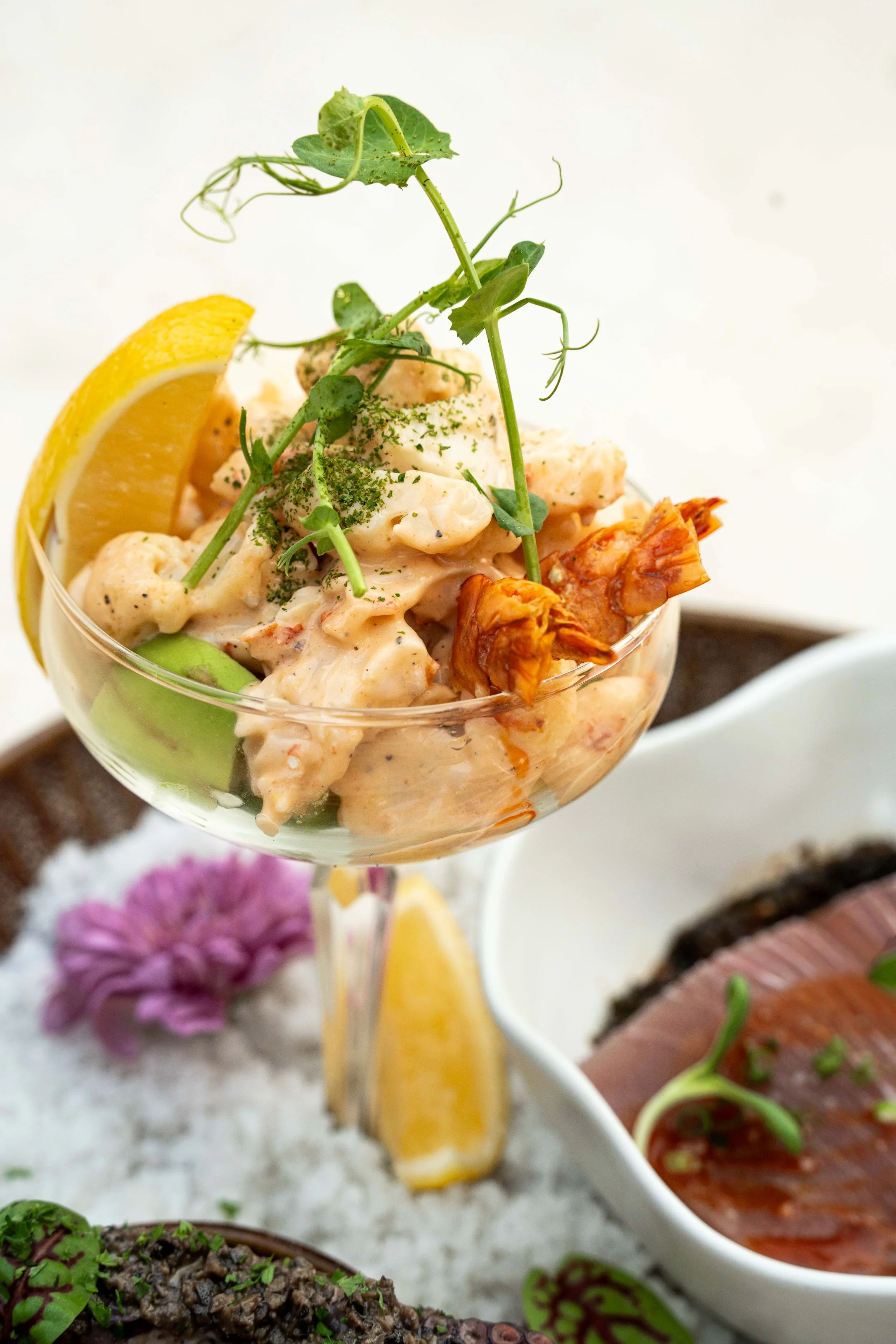 Close-up of a seafood dish in a glass, garnished with lemon wedges, herbs, and microgreens, with additional seafood dishes and flowers in the background.