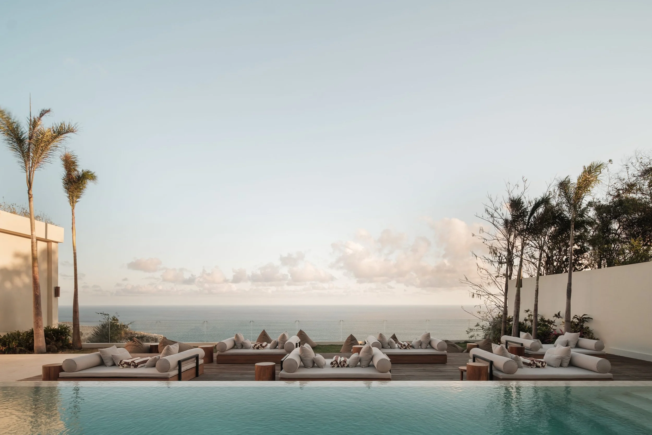 Outdoor poolside lounge area with cushioned seating, palm trees, and a view of the ocean in the distance, during sunset.