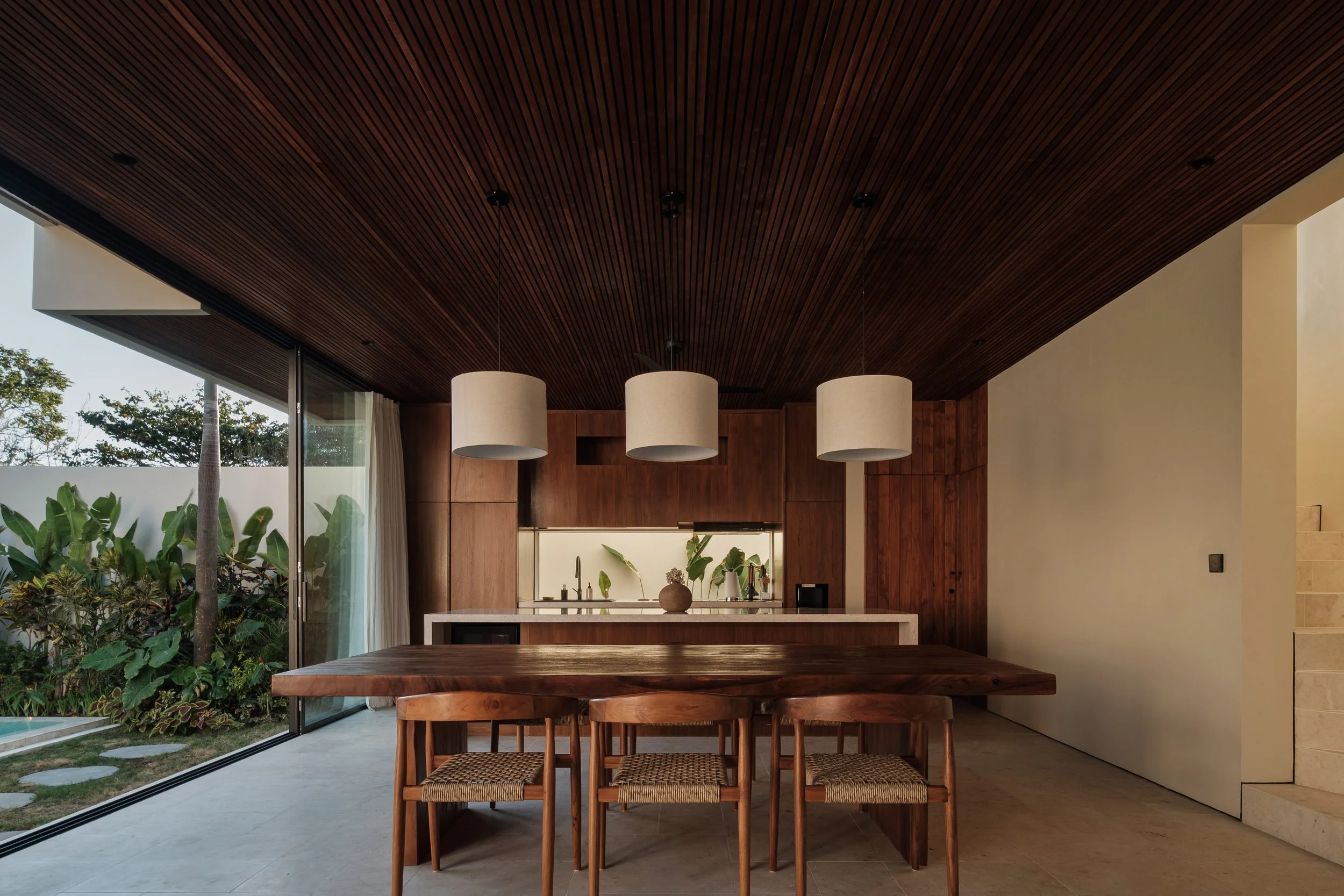 Modern dining area with wooden table and stools, open kitchen, dark wooden ceiling, and sliding glass doors to outdoor garden with plants and trees.