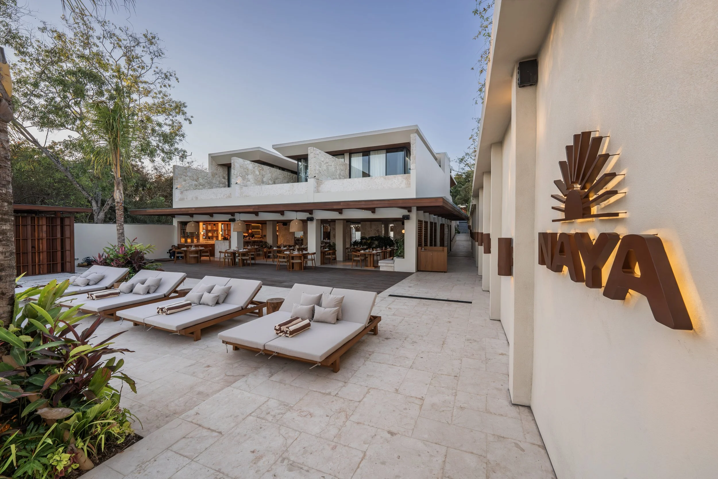 Outdoor patio area of a modern building with lounge chairs, tables, trees, and a bar area, featuring a two-story structure with large windows and a decorative sunburst sign on the wall.