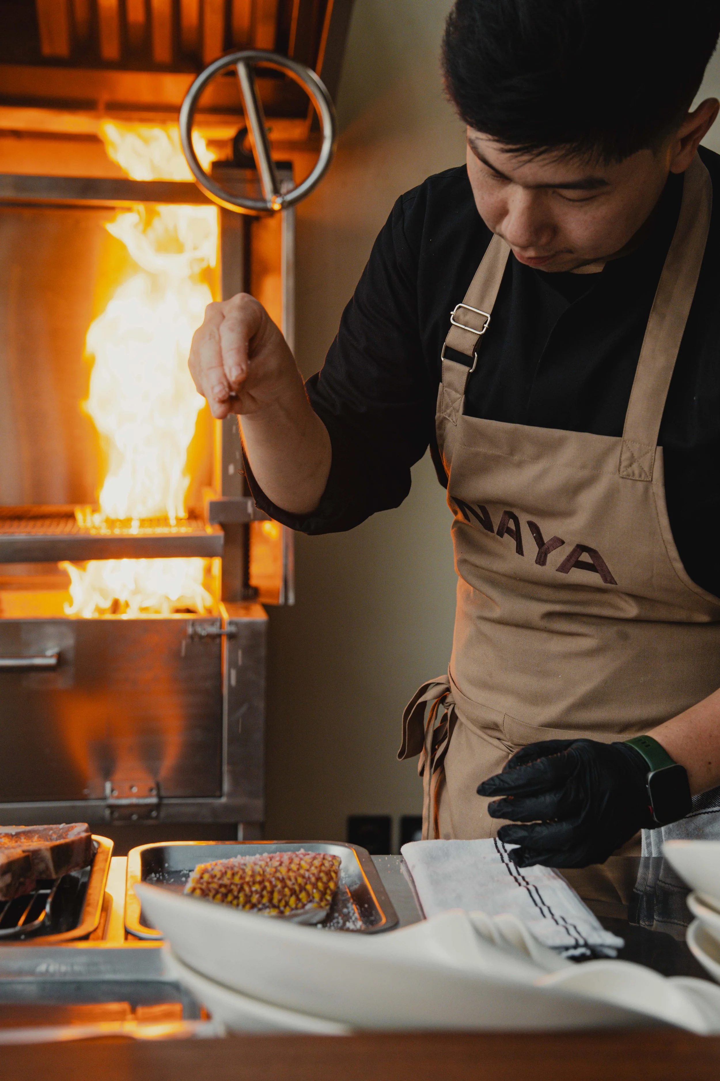 A person wearing a black shirt, beige apron, and black gloves preparing food in a professional kitchen. Fire is visible in the background, suggesting cooking or a culinary demonstration.