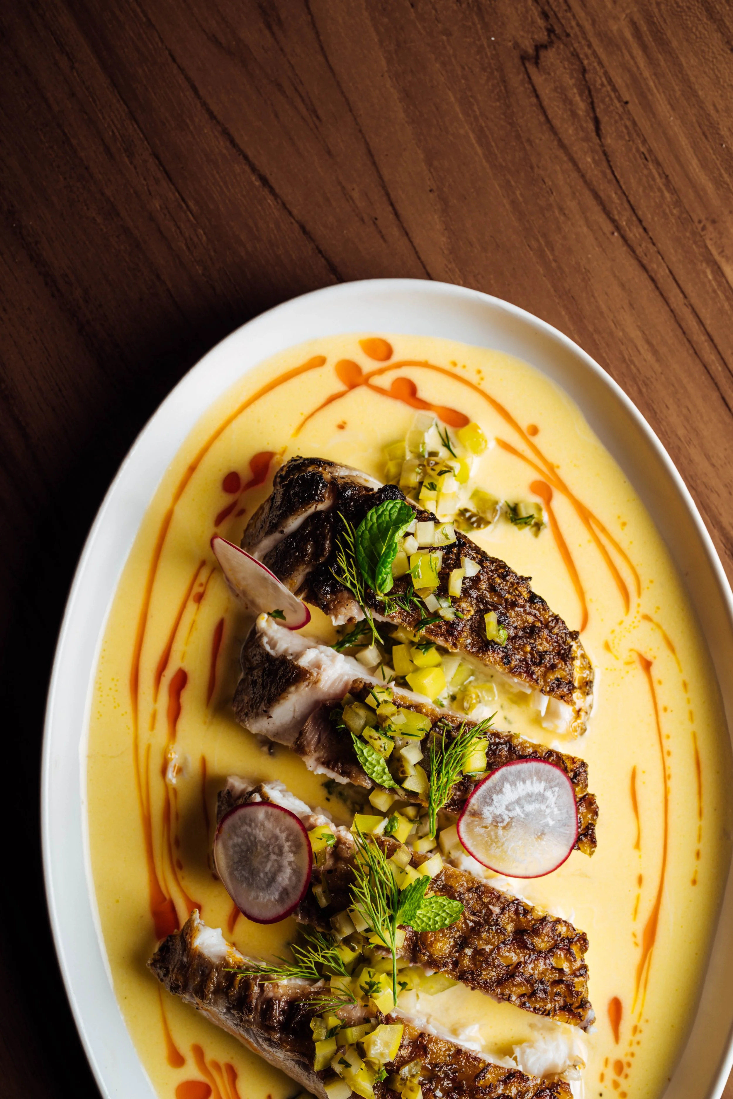 A plate of cooked fish served on a bed of yellow sauce, garnished with radish slices, chopped green onions, fresh herbs, and drizzled with orange sauce.