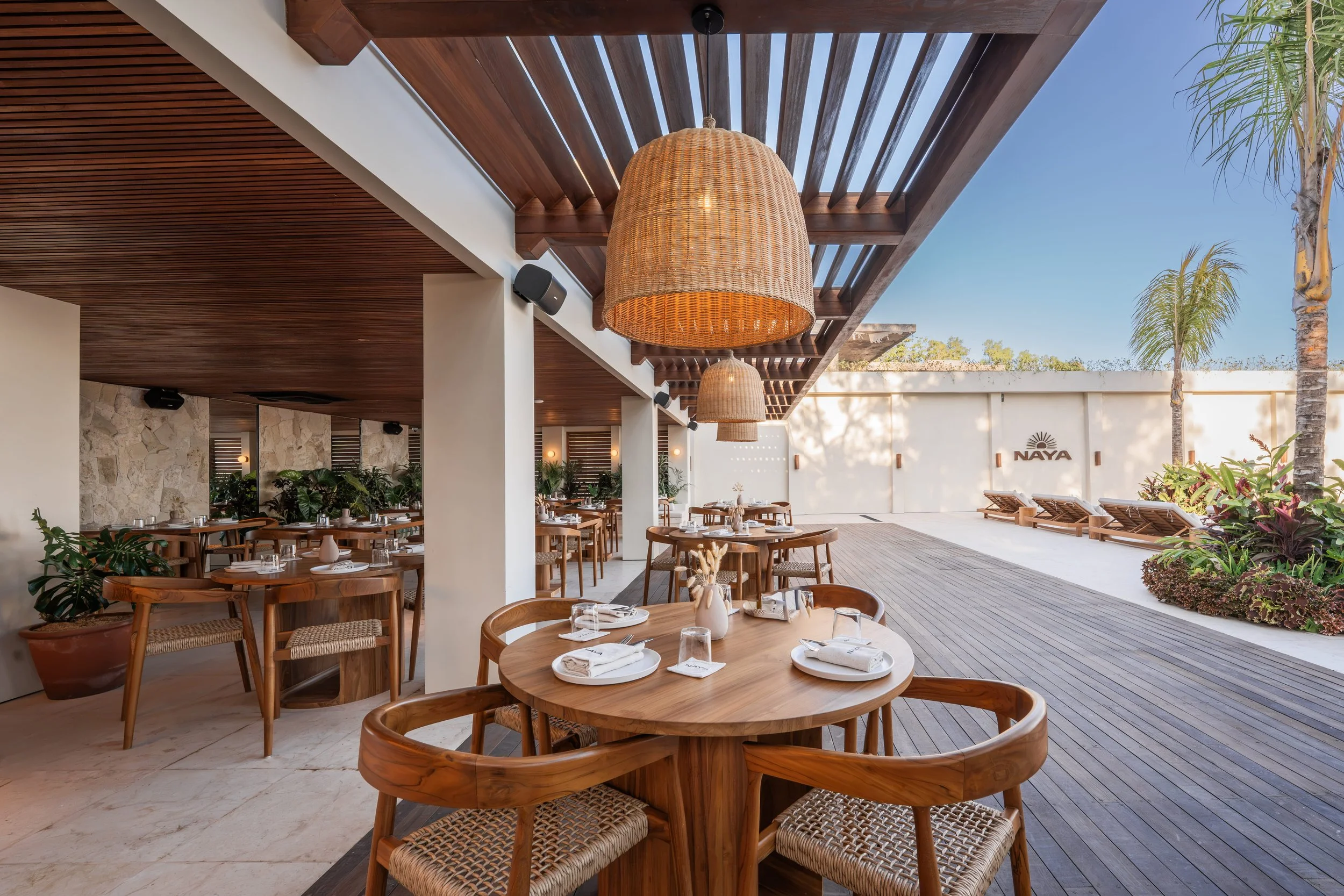 Outdoor dining area with round wooden tables set with plates, glasses, and napkins, surrounded by wooden chairs featuring woven seats, under large wicker pendant lights. Partially covered with wooden slats, open to a clear blue sky and landscaped wit