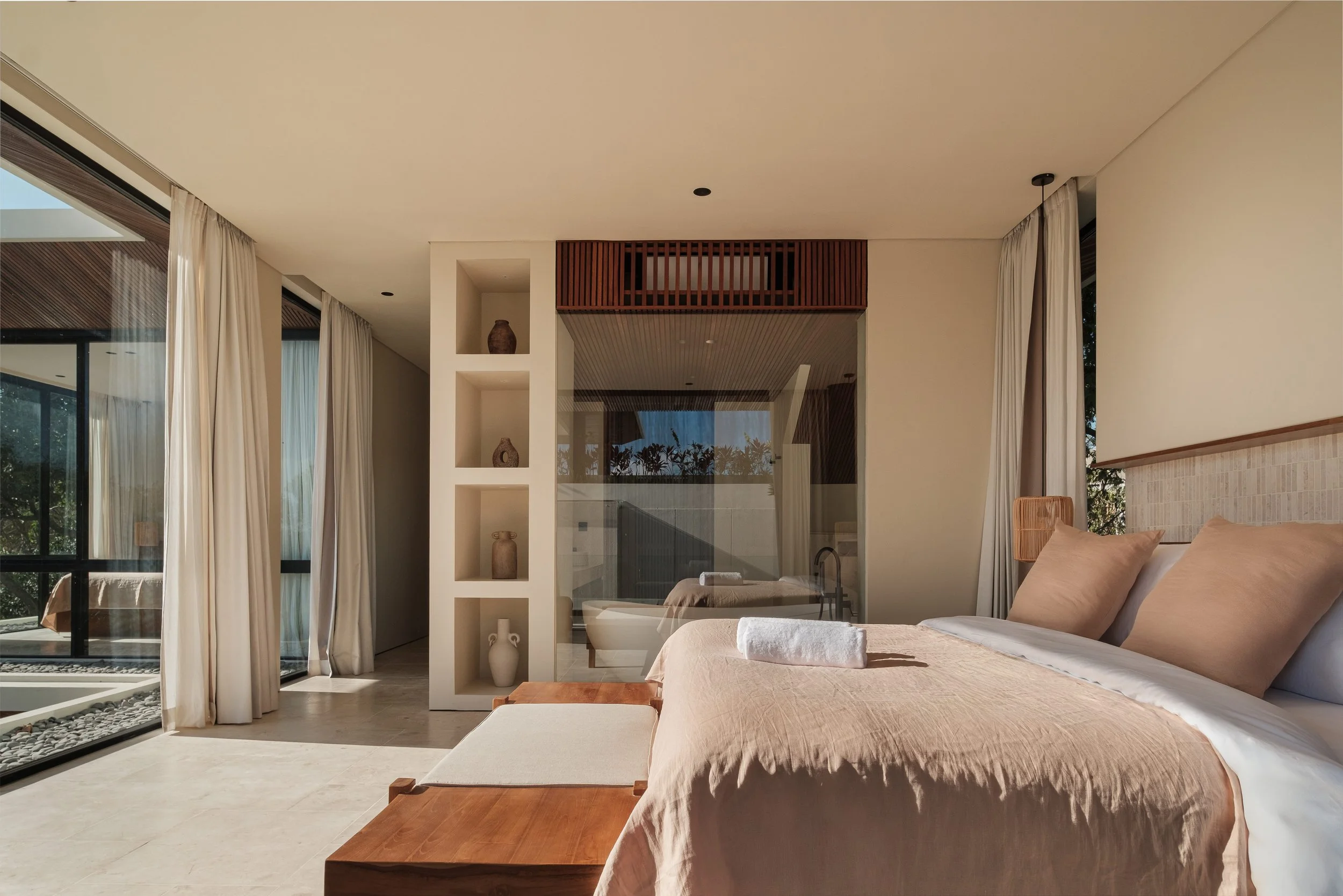 Modern bedroom with beige bedding, pillows, wooden furniture, floor-to-ceiling windows with white curtains, and decorative pottery in a built-in shelf.