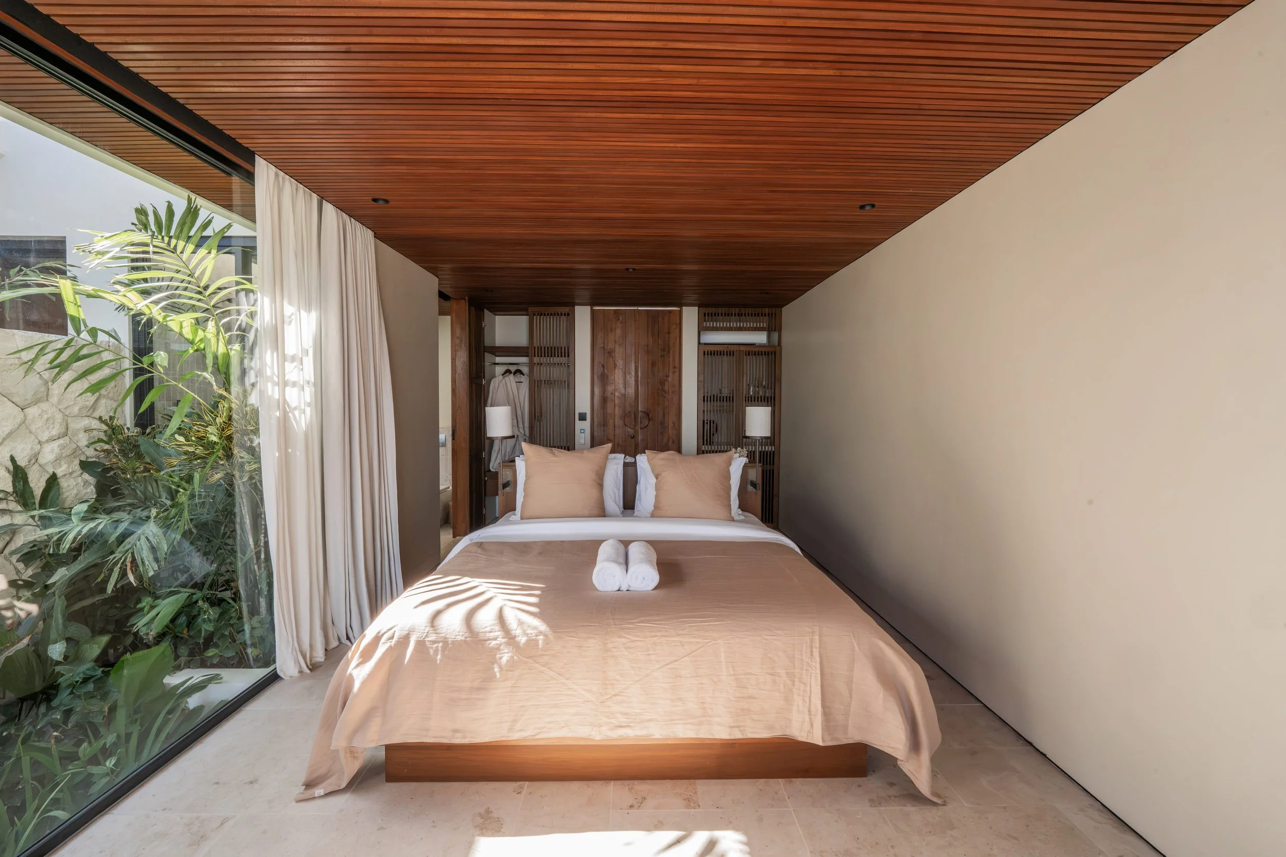 A modern bedroom with a wooden ceiling, beige bed, white and beige pillows, and a large sliding glass door with green plants outside.