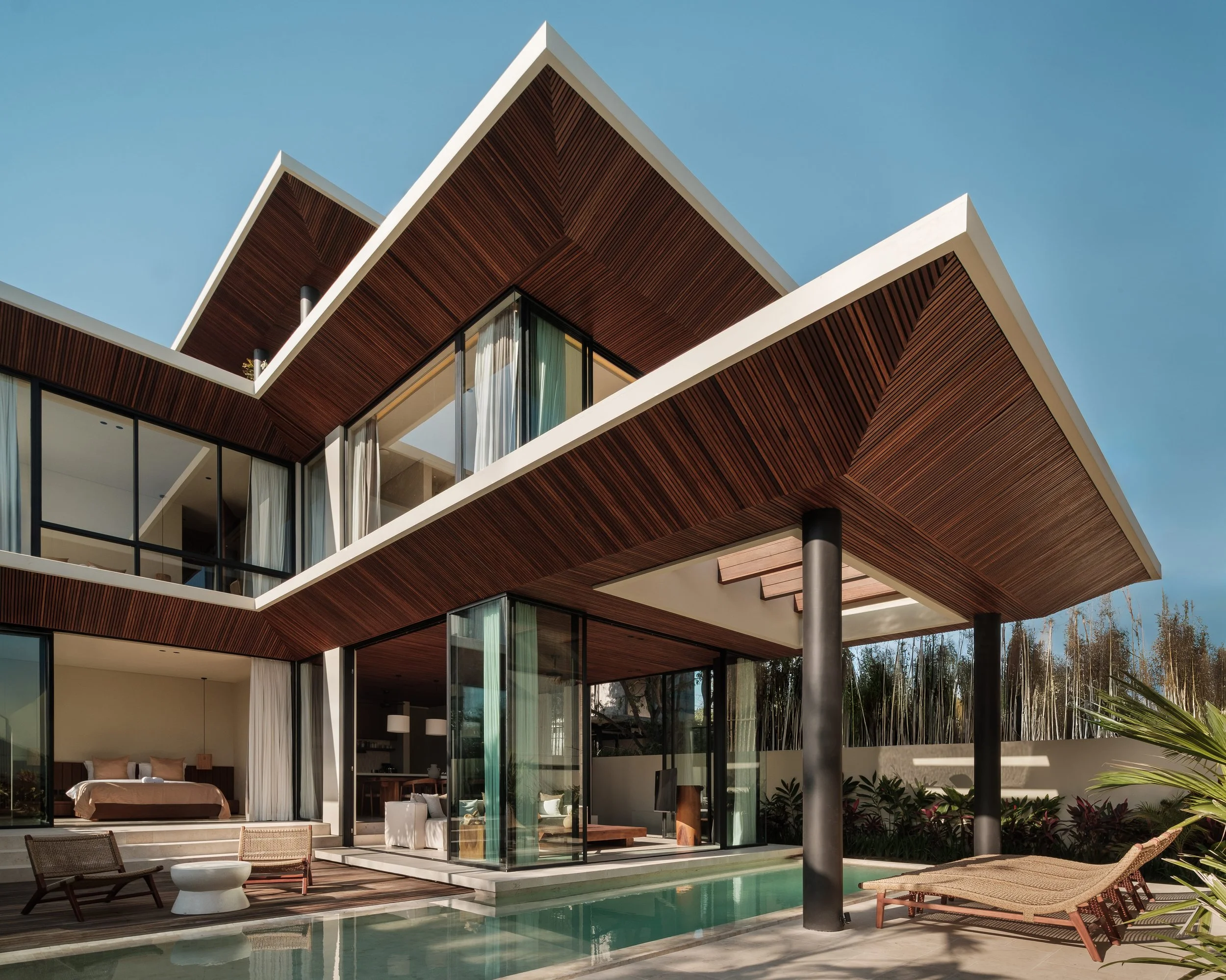 Modern house with large glass windows, wooden accents, and a swimming pool in the foreground, under a clear blue sky.