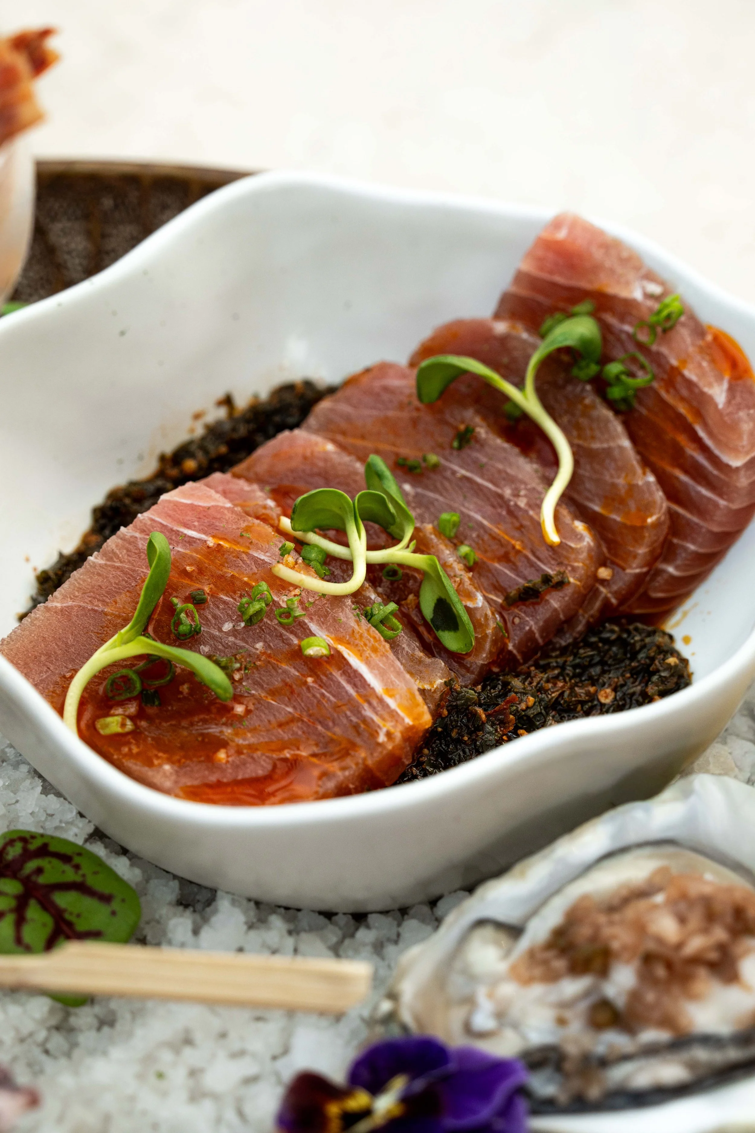 Slices of raw tuna sashimi garnished with microgreens in a white dish, with a background of various sushi rolls and flowers on a bed of salt.