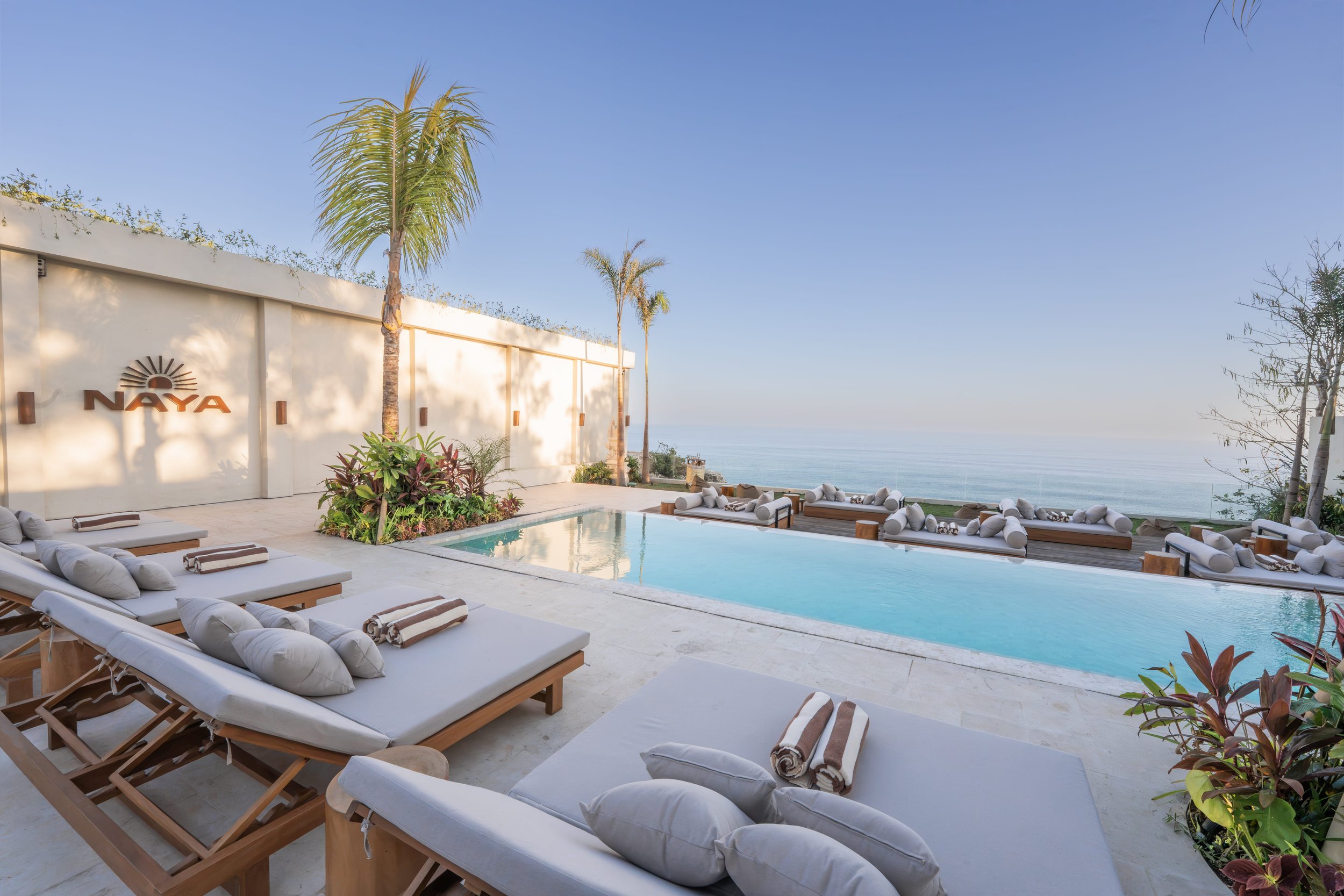 Luxury outdoor pool area with lounge chairs, rolled towels, and tropical plants, overlooking the ocean under a clear blue sky at Naya resort.