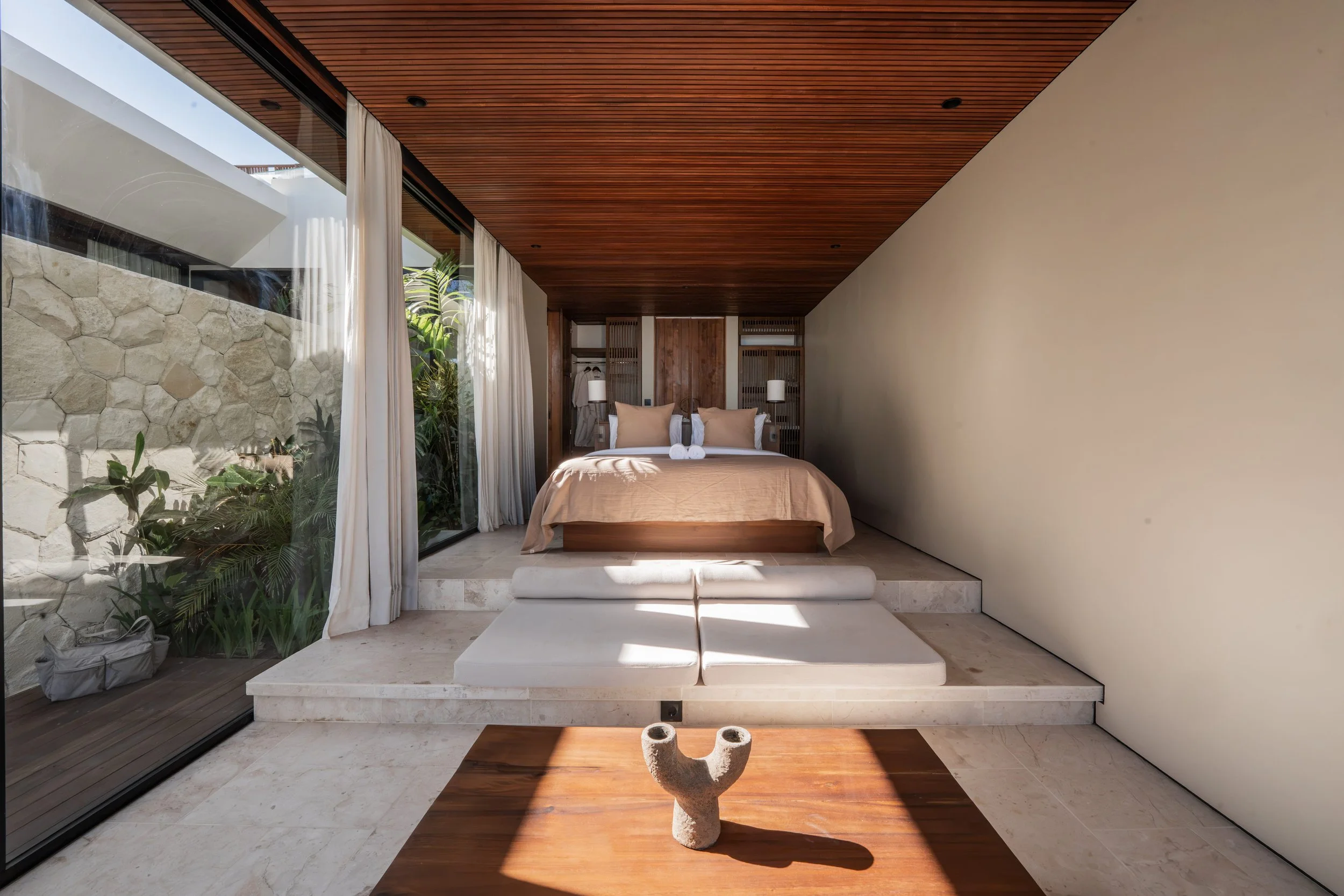 Modern bedroom with large glass sliding door, wooden ceiling, stone and beige walls, and minimalist decor including a bed with beige pillows, white towels, a wooden coffee table with a decorative sculpture, and green plants outside.