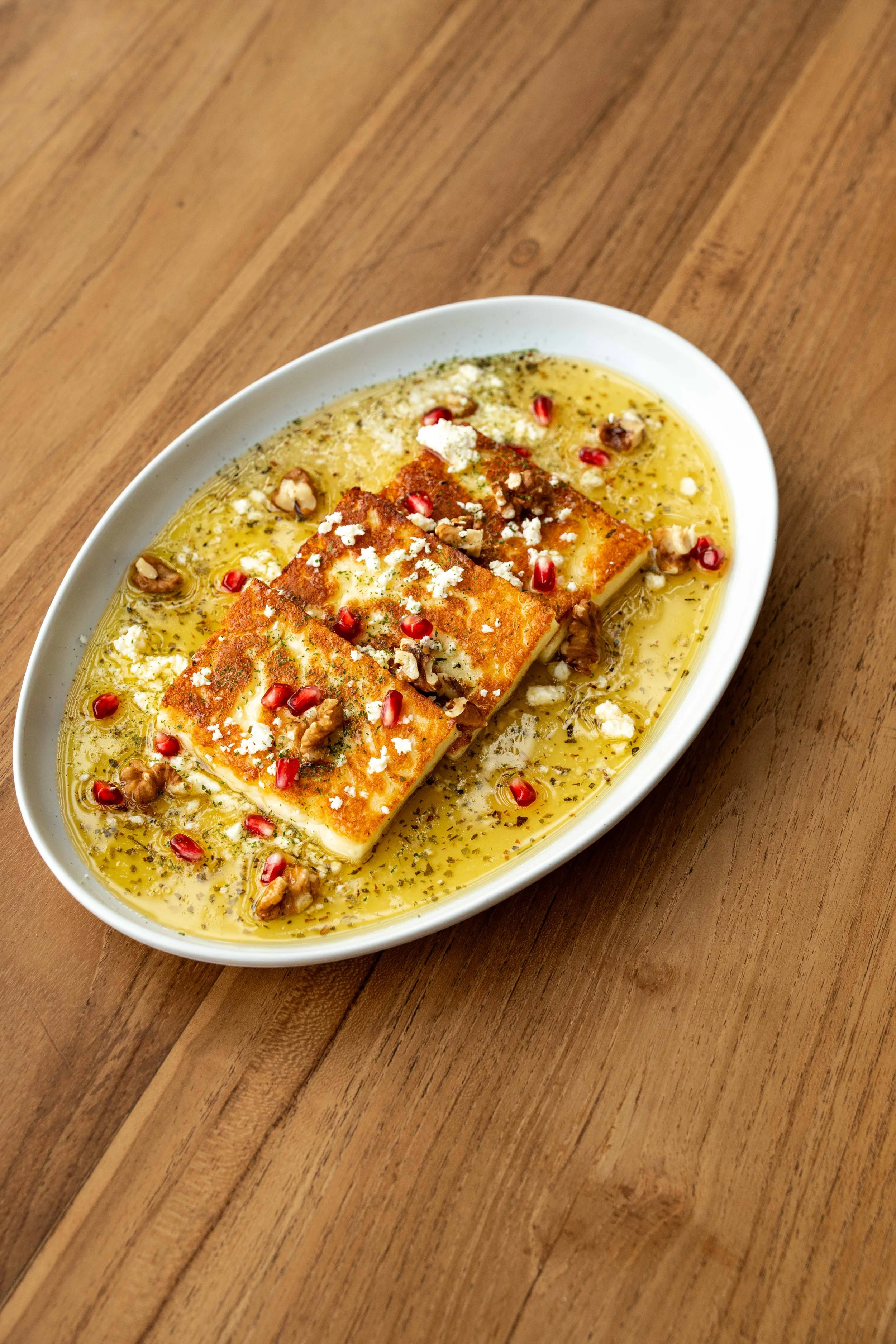 A dish of fried cheese topped with pomegranate seeds, chopped nuts, and herbs in oil or sauce on a white oval plate on a wooden surface.
