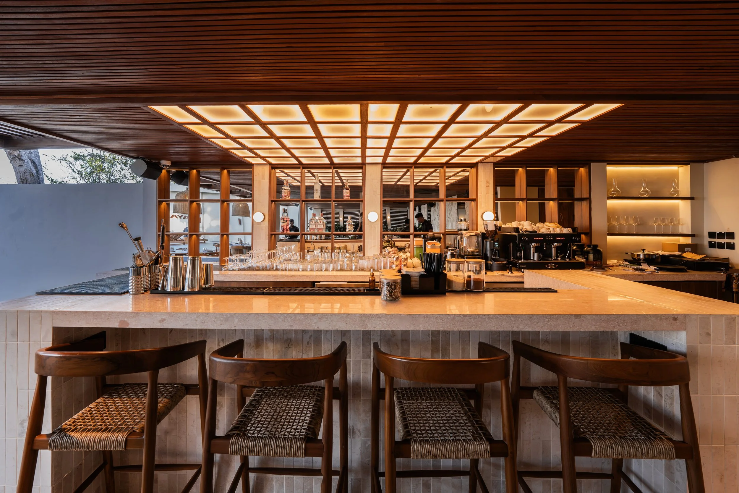 Modern restaurant or bar interior with a bar counter, bar stools, wooden ceiling with backlit panels, and shelves with liquor bottles and glassware.