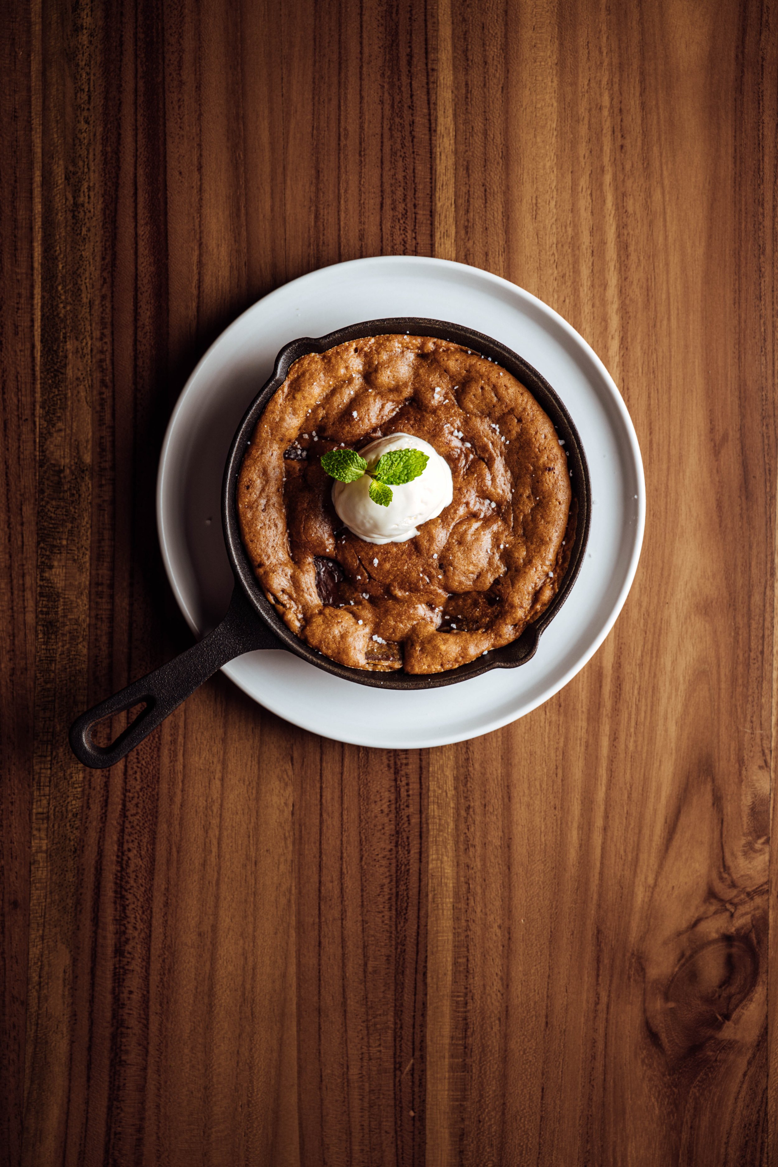 A dessert in a small cast iron skillet with a scoop of ice cream and a sprig of mint on top, served on a white plate on a wooden table.
