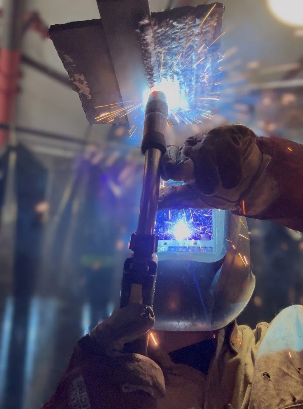 A person welding metal with sparks flying in an industrial workshop, wearing protective gear including gloves and a welding helmet.
