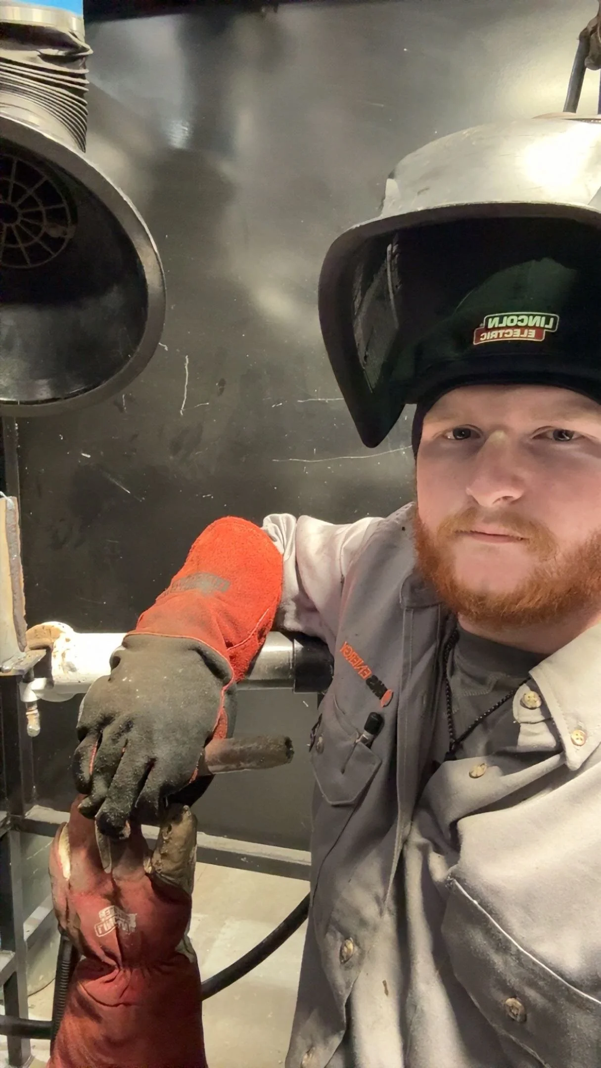 A man wearing a welding helmet on his head, gray work shirt, black gloves, and a gray jumpsuit, taking a selfie in an industrial workshop.