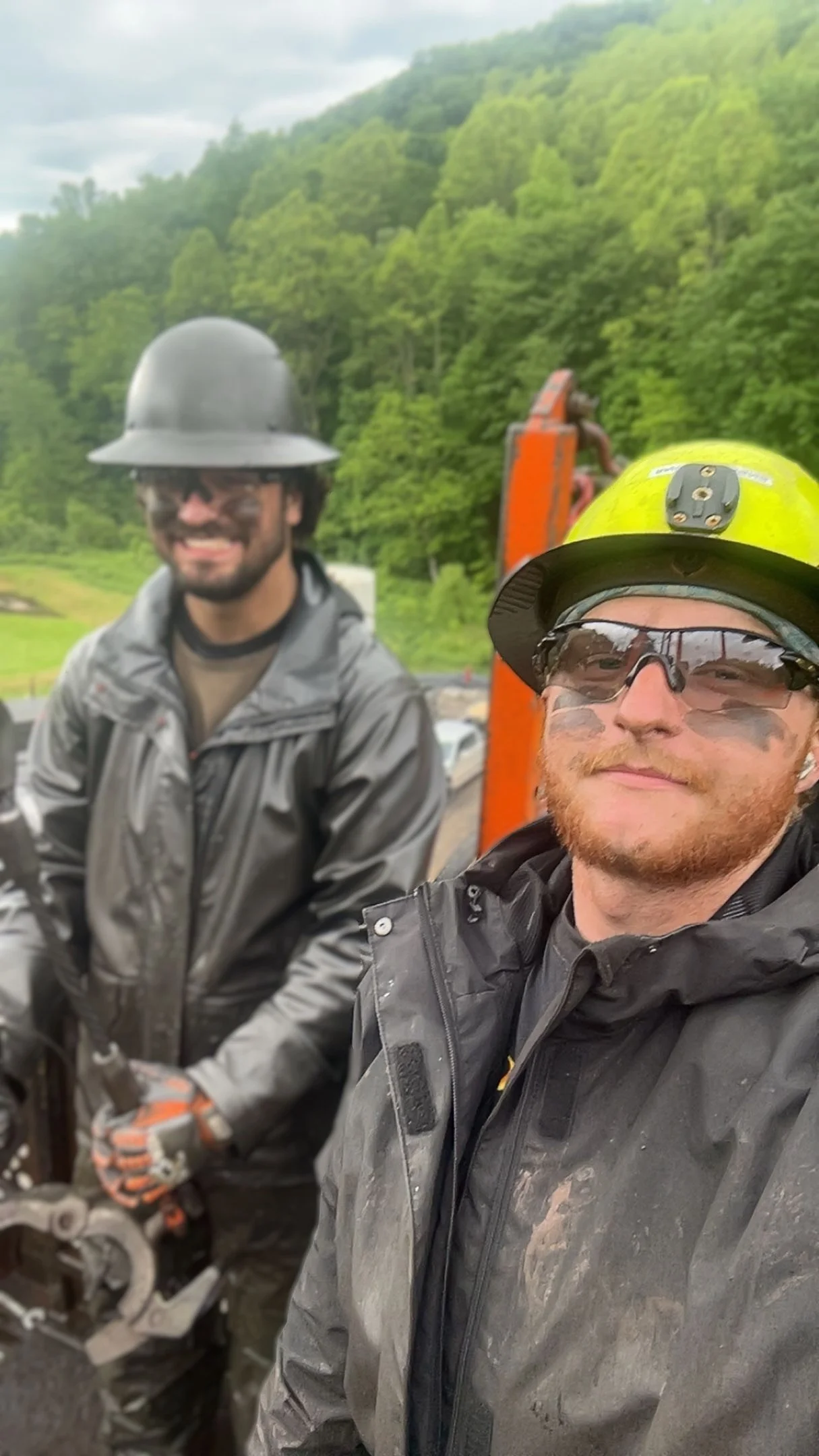 Two men in safety gear, helmets, and goggles standing on a construction or mining site with green trees and hills in the background.