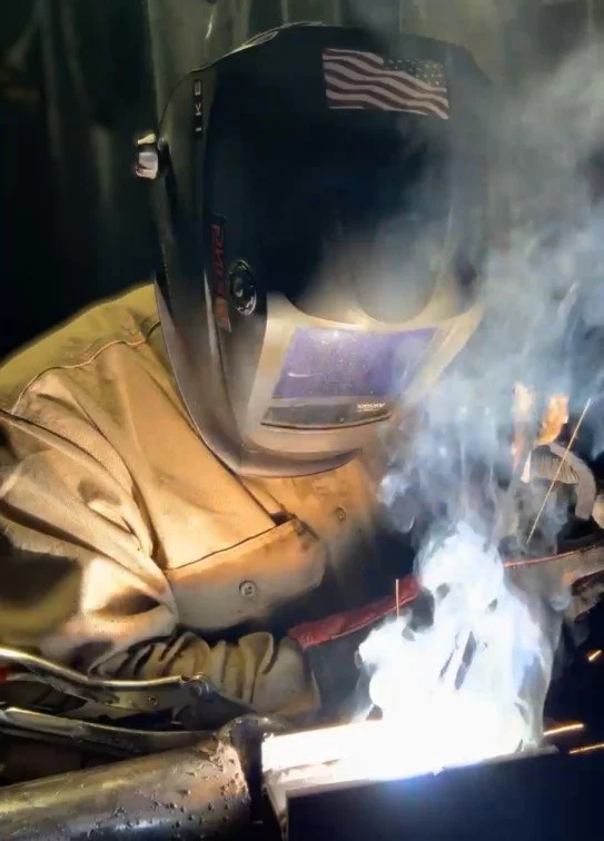 A person welding in a dark environment, wearing a protective welding helmet and tan jacket, with sparks and smoke around.