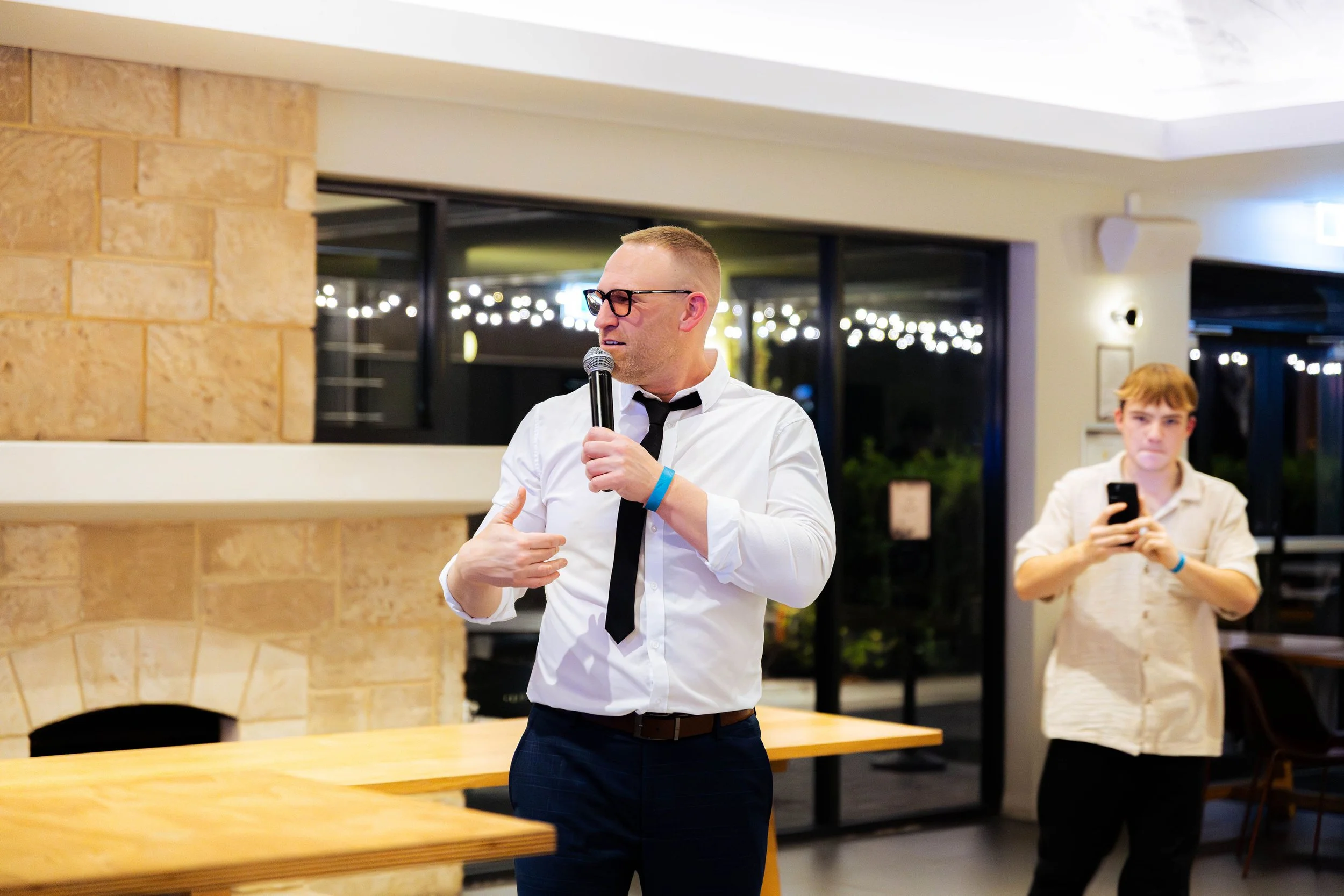A man in a white shirt, black tie, and glasses is speaking into a microphone while gesturing with his hand. A young man in a light-colored shirt is taking a photo with his phone in the background. The setting appears to be indoors in a modern, well-lit room with large windows and a stone fireplace.