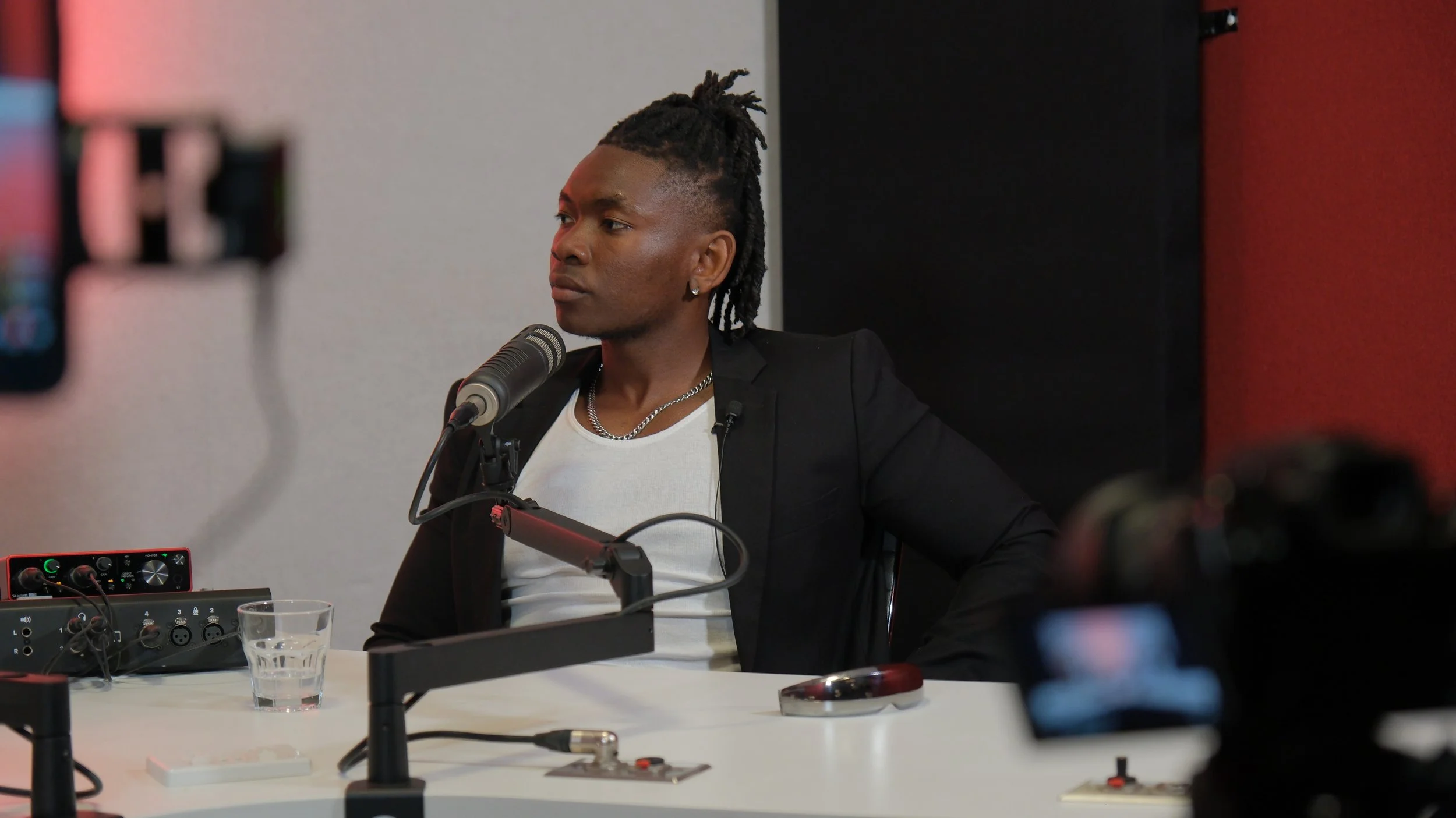 A man with dreadlocks and earrings is sitting at a radio or podcast studio table, speaking into a microphone with recording equipment around him.