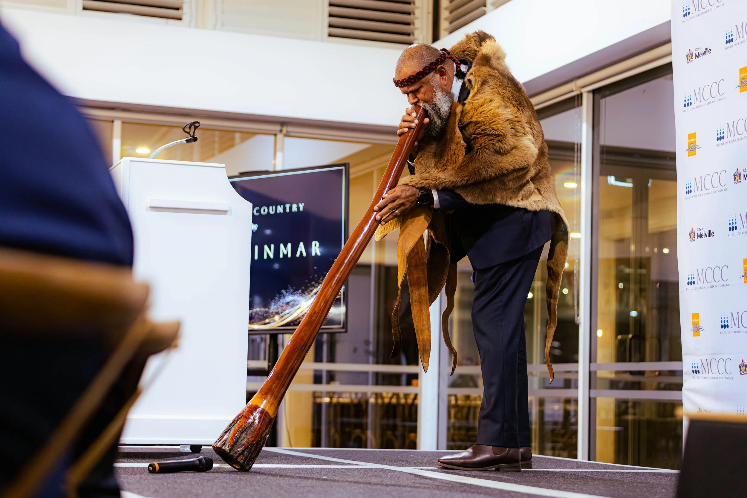 An Indigenous man dressed in traditional attire plays a wooden didgeridoo at an indoor event. He is wearing a fur shawl, a red and black headband, and black pants. There is a banner with logos in the background.