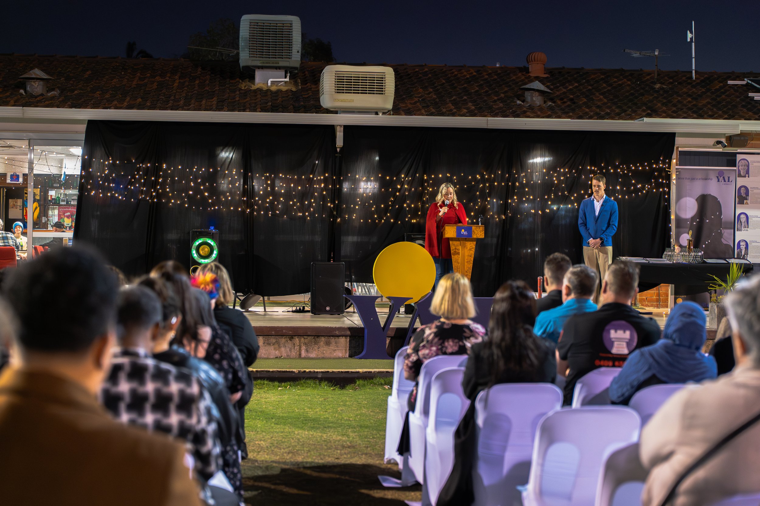 Nighttime outdoor event with a stage, where a woman in red and a man in blue are speaking, audience seated on chairs, black curtain with string lights behind the speakers, and people attending.