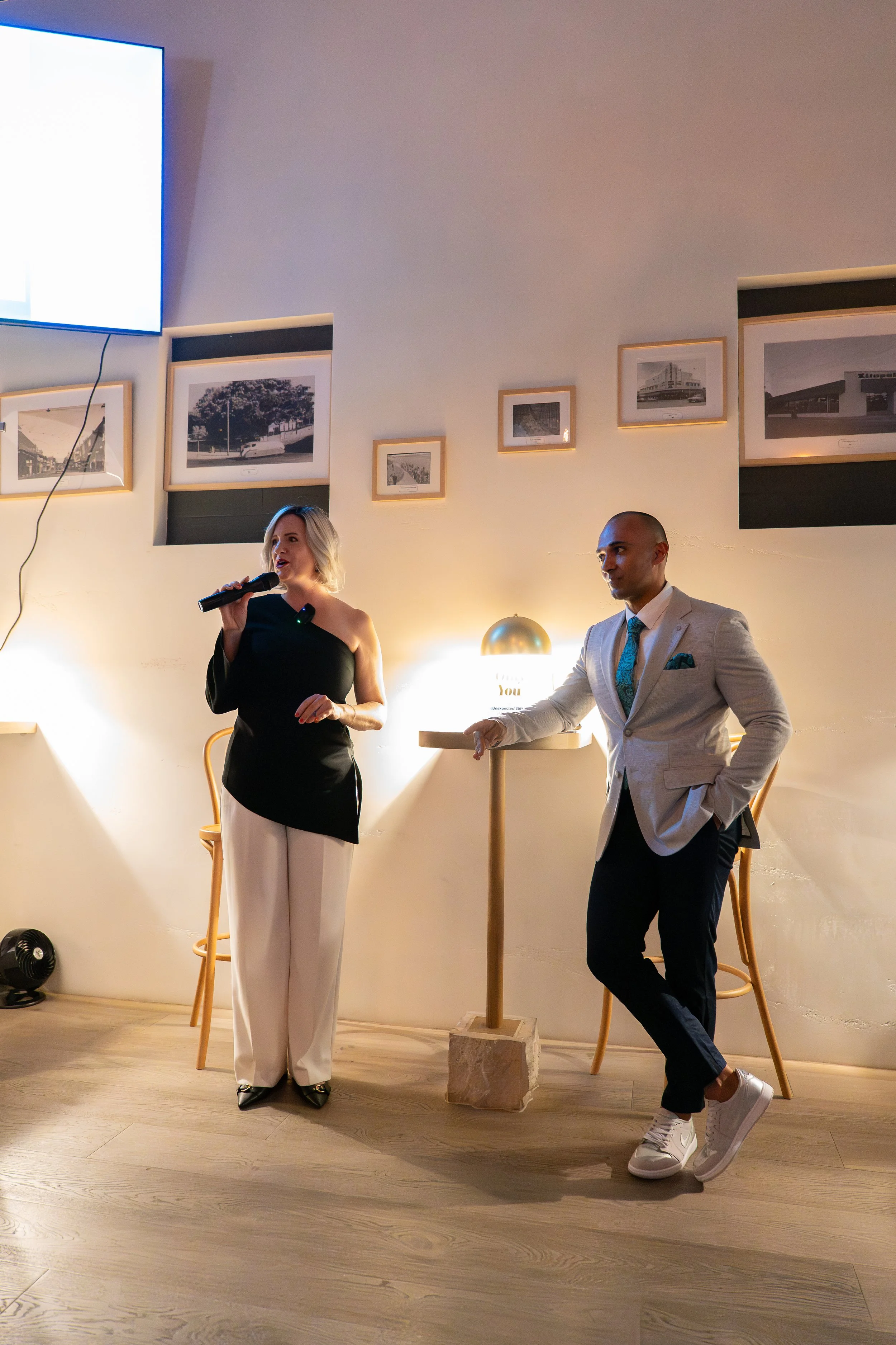 A woman in black top and white pants holding a microphone standing next to a man in a light-colored suit, both inside a room with framed pictures on the wall. The man is leaning on a high table, and there are chairs and a lamp in the background.