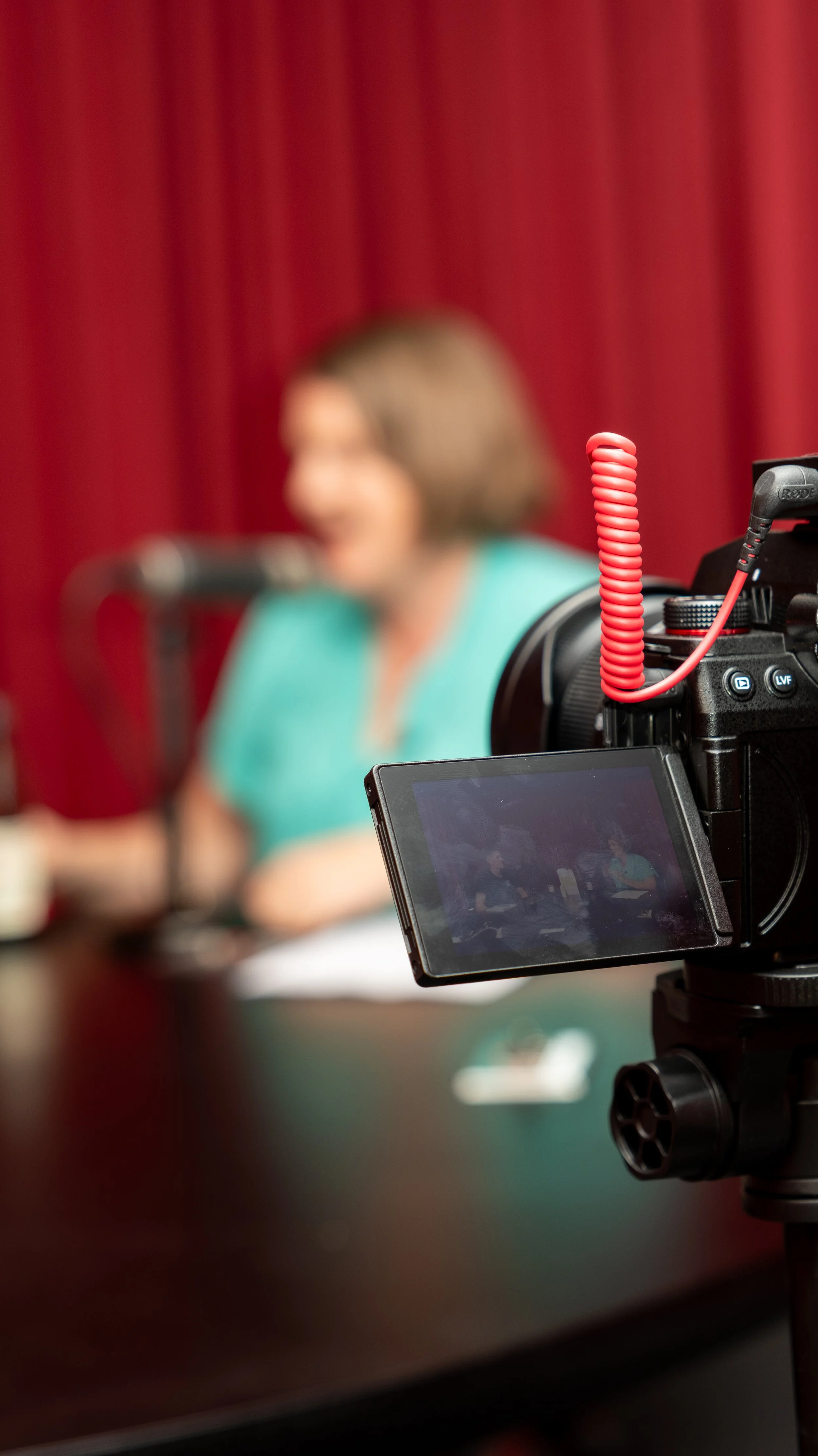 A camera focused on a woman speaking into a microphone, with a red curtain backdrop. The woman is wearing a teal top and is slightly out of focus.