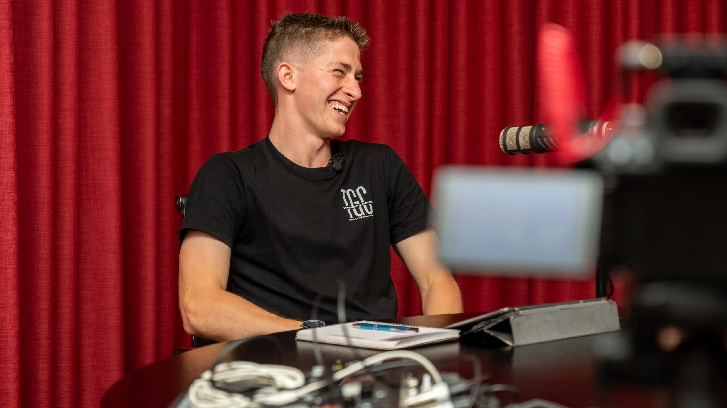 A young man with short hair smiling and laughing while sitting at a table with a notebook, pen, and electronic devices, in front of a red curtain background.