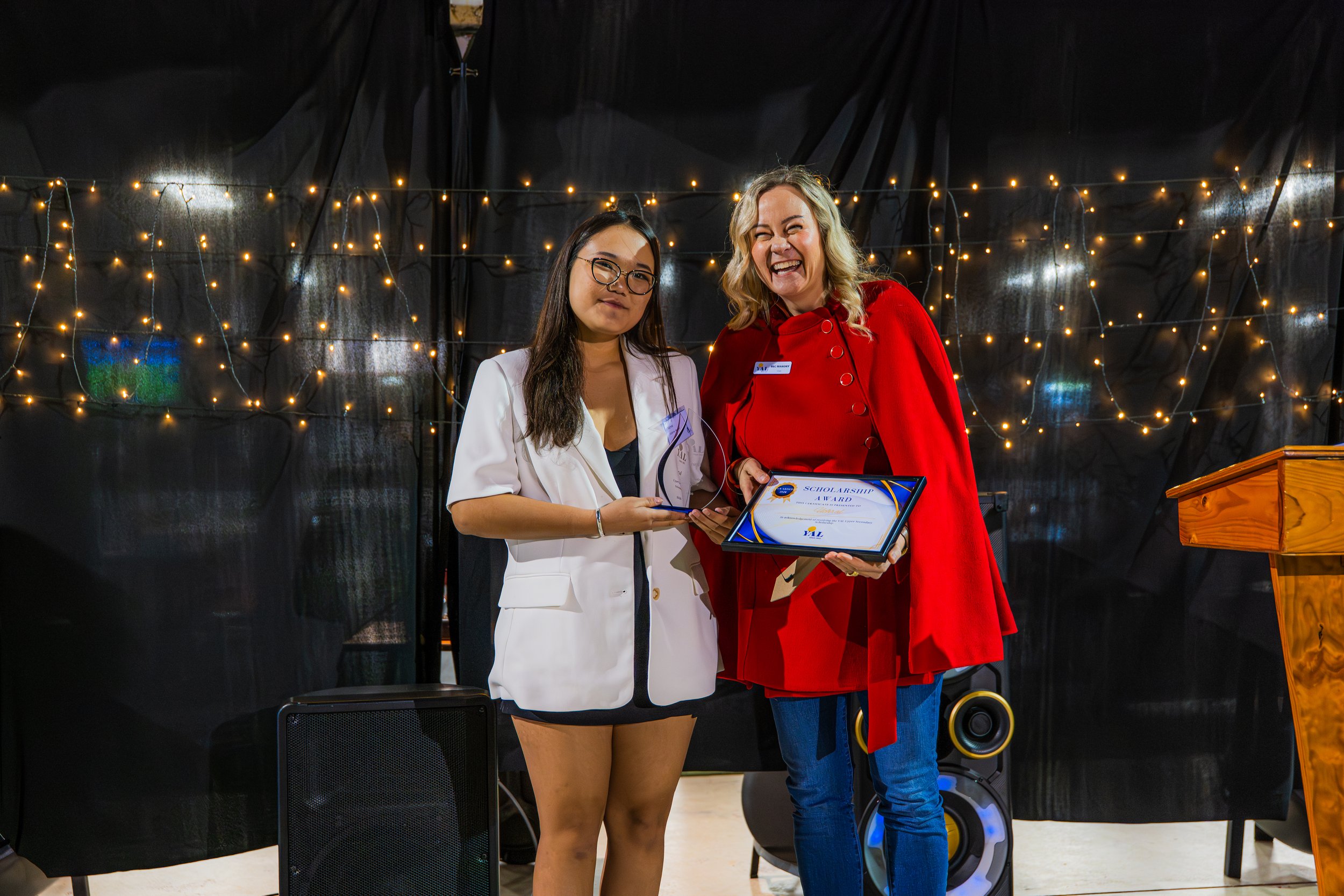 Two women standing on a stage with a black curtain and string lights behind them, one holding a scholarship award, and both smiling.