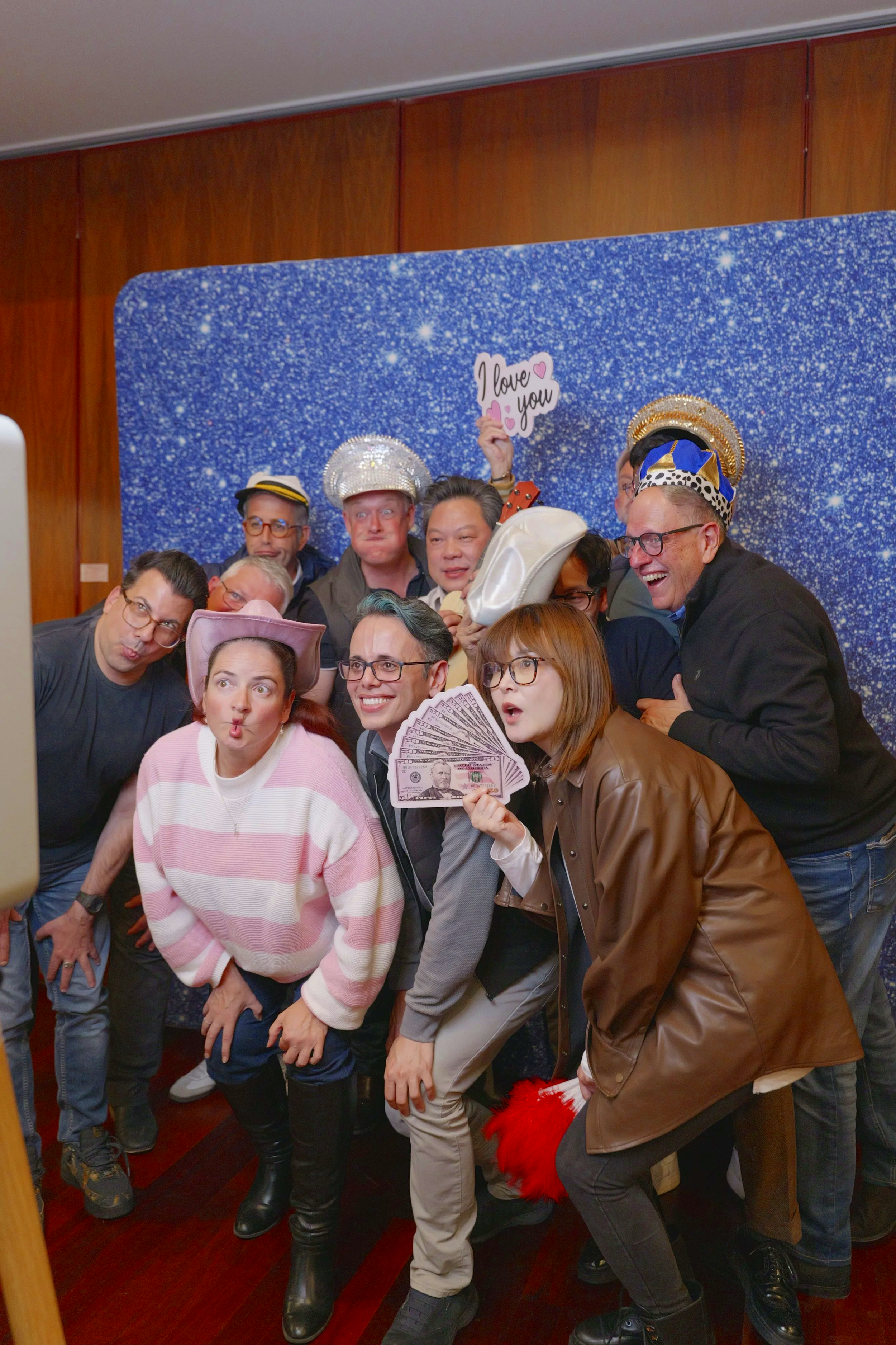 Group of people dressed in humorous costumes posing for a photo in front of a sparkly blue backdrop, some wearing hats and holding props.