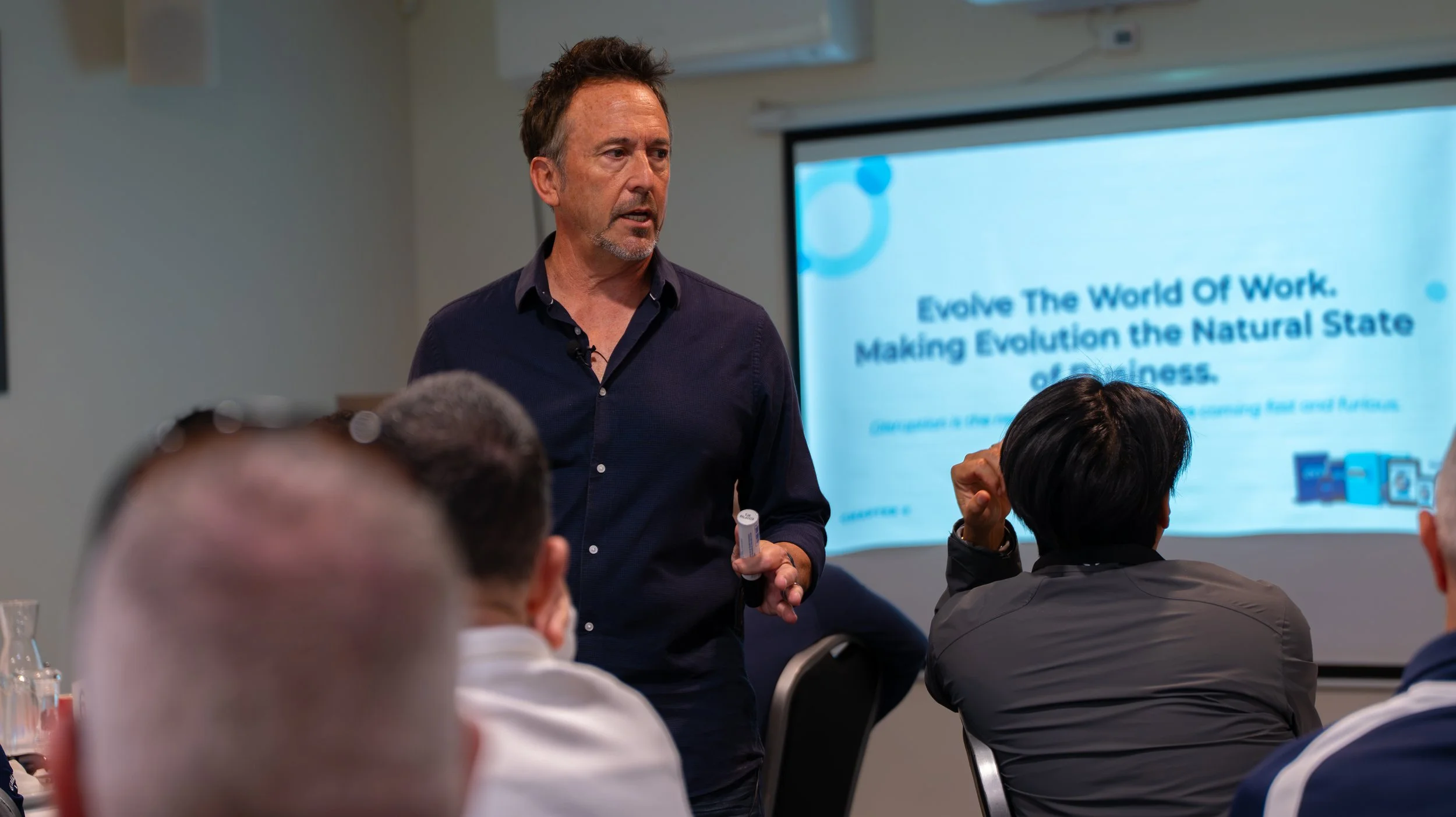 A man in a dark shirt giving a presentation to a group of people in a conference room, with a slide titled 'Evolve The World Of Work. Making Evolution the Natural State of Business' projected on the wall behind him.