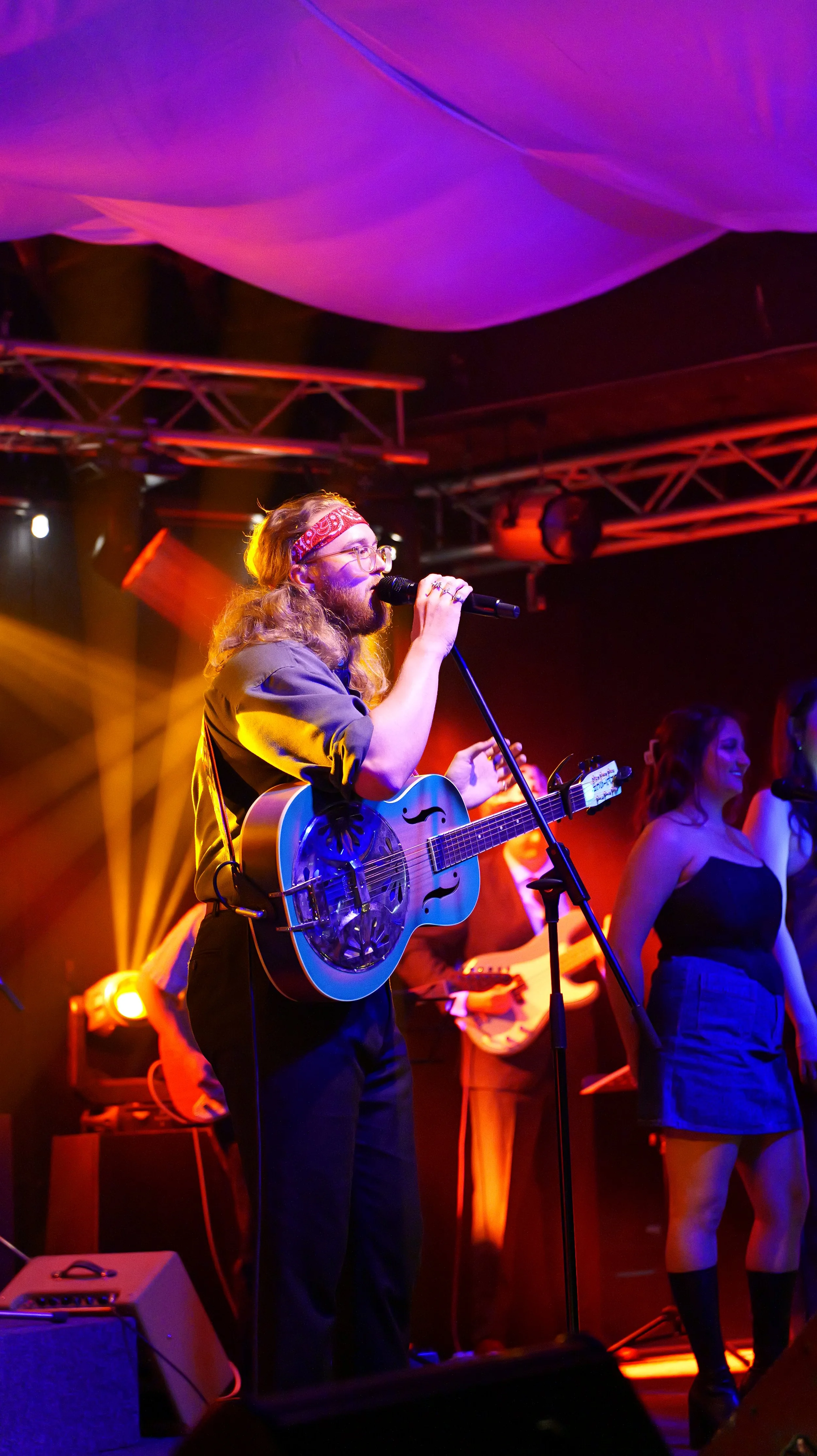 A male musician with long hair, glasses, and a red bandana singing into a microphone and playing an acoustic guitar on stage with colorful lighting, surrounded by backup singers and musicians.