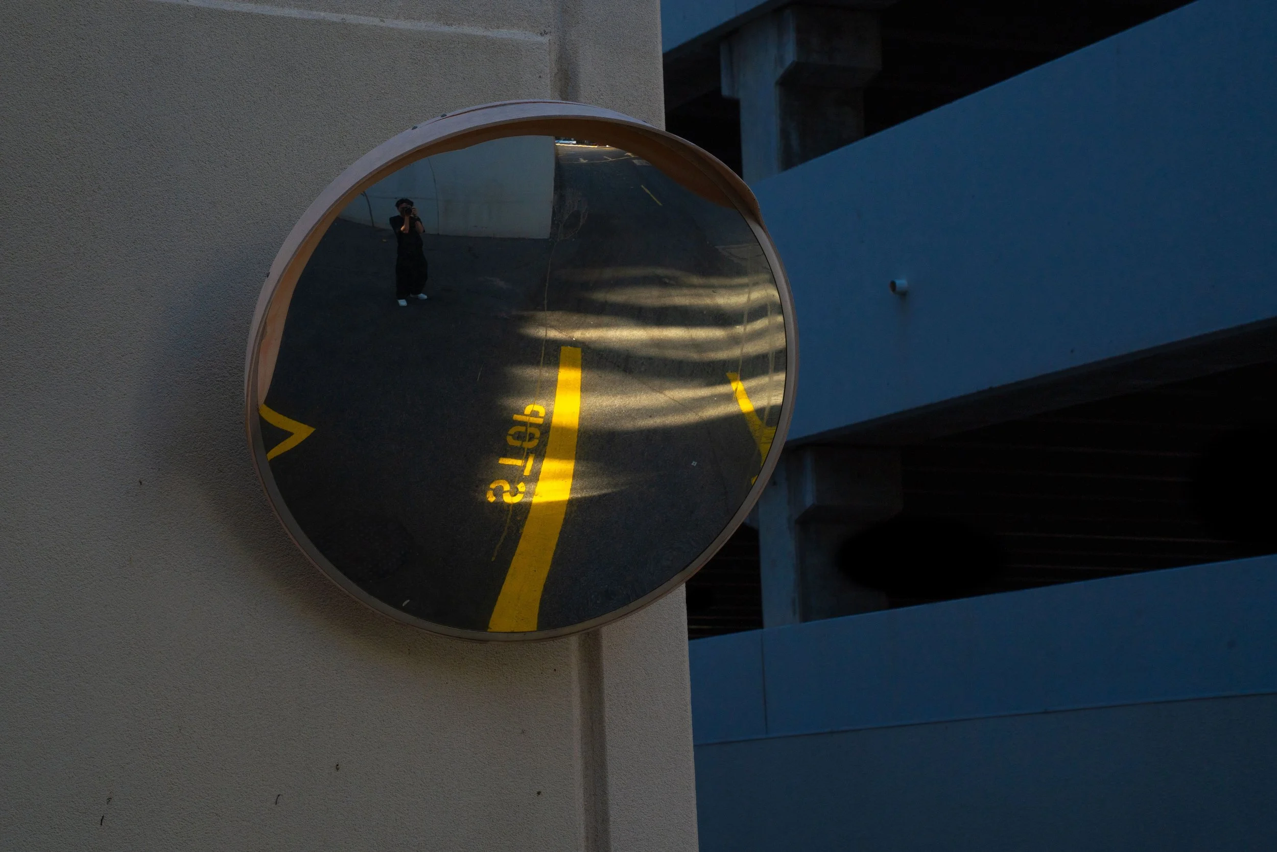 A round convex mirror mounted on a wall reflecting a parking garage with yellow painted parking lines, a person taking a photo, and shadows.