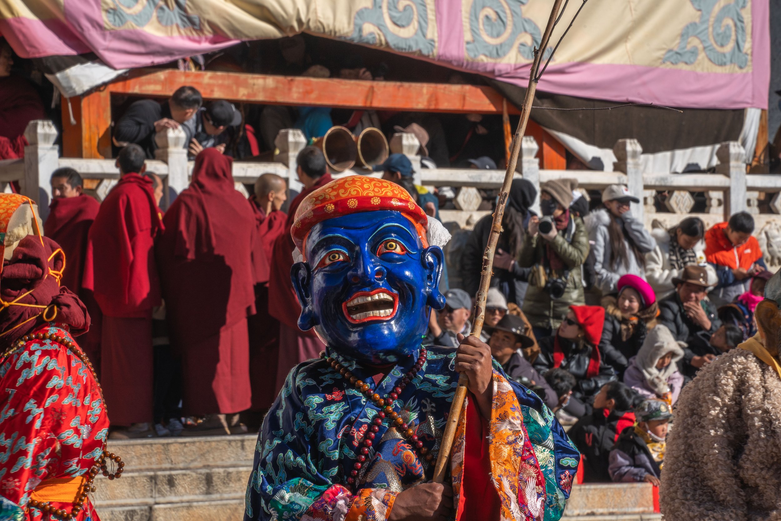 A person in traditional attire wearing a bright blue mask and holding a staff during a cultural festival, with spectators watching in the background.