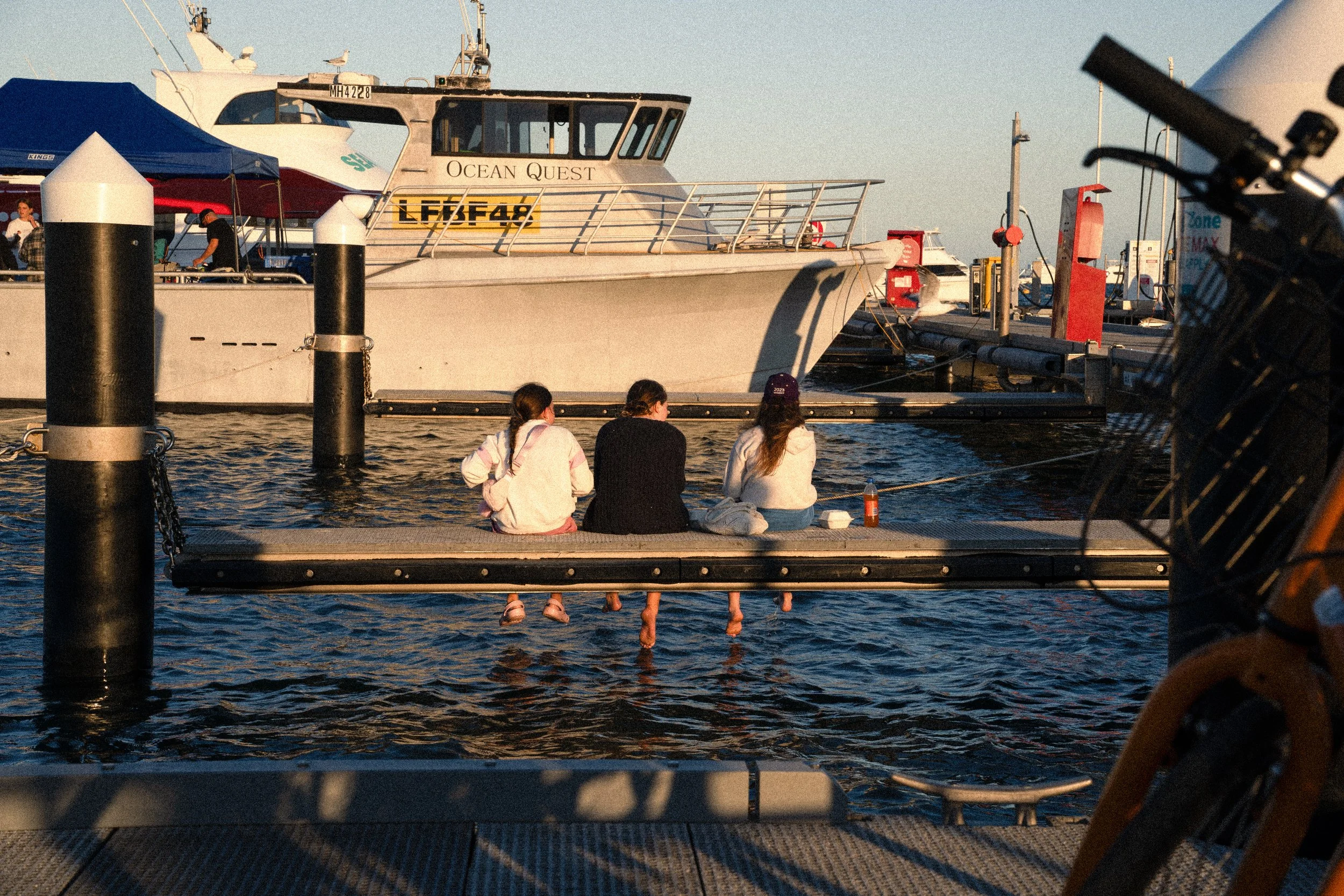 Three people sitting on a dock by the water, with boats and a yacht nearby, during sunset.