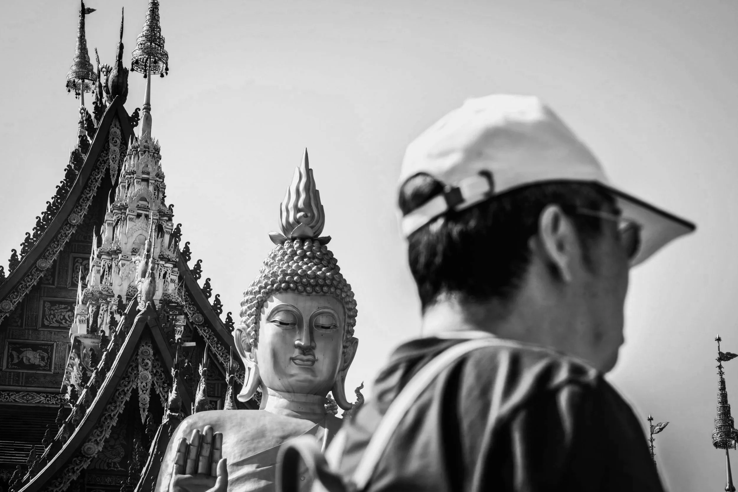 A person wearing a white cap and sunglasses in front of a large golden Buddha statue and ornate temple in Thailand.