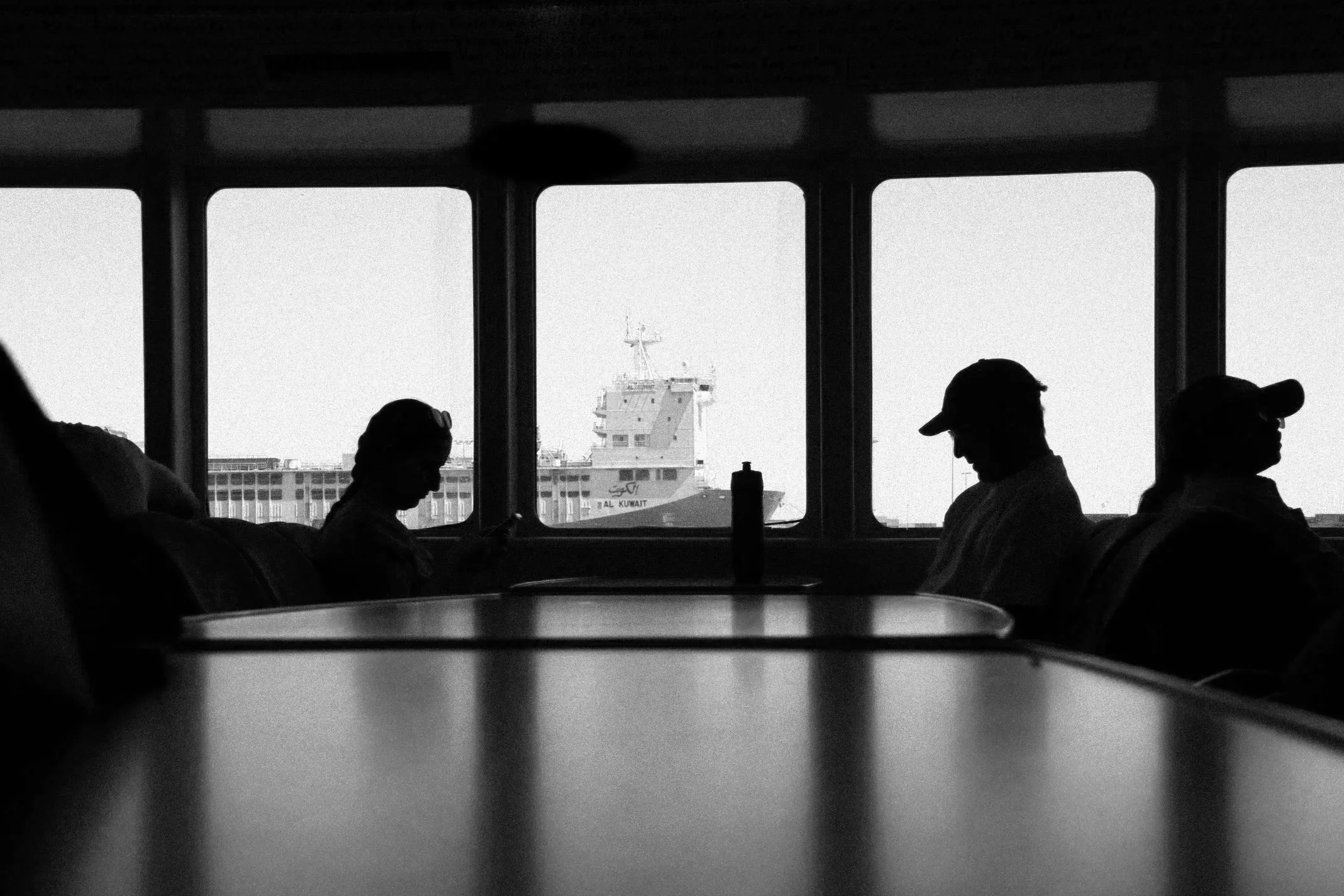 Silhouetted passengers sitting inside an airport waiting area or waiting lounge, with a large window showing a ship anchored outside, labeled 'Al Kuwait,' against a clear blue sky.