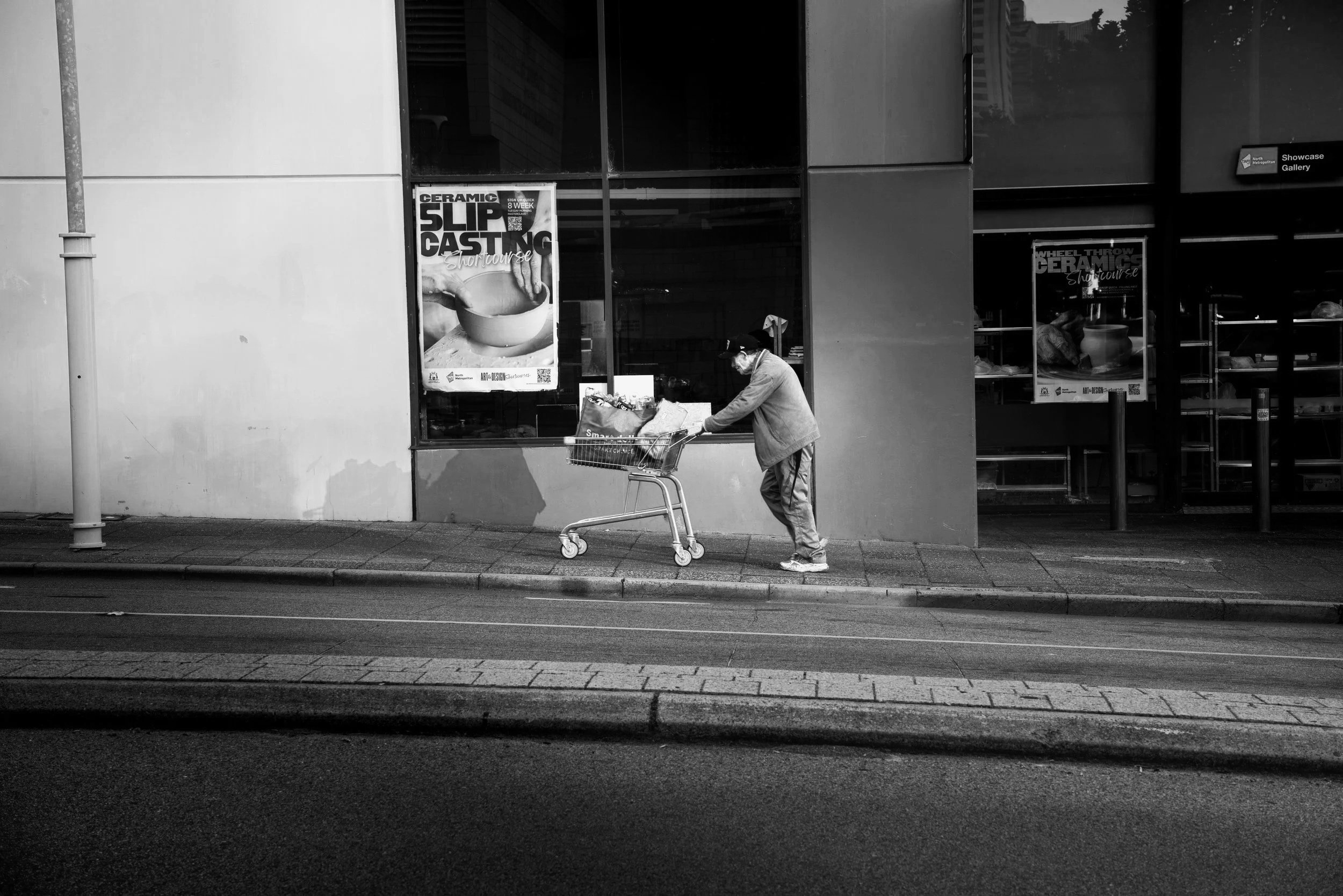 A man wearing a brown jacket and cap pushing a shopping cart on a city sidewalk in front of a glass building