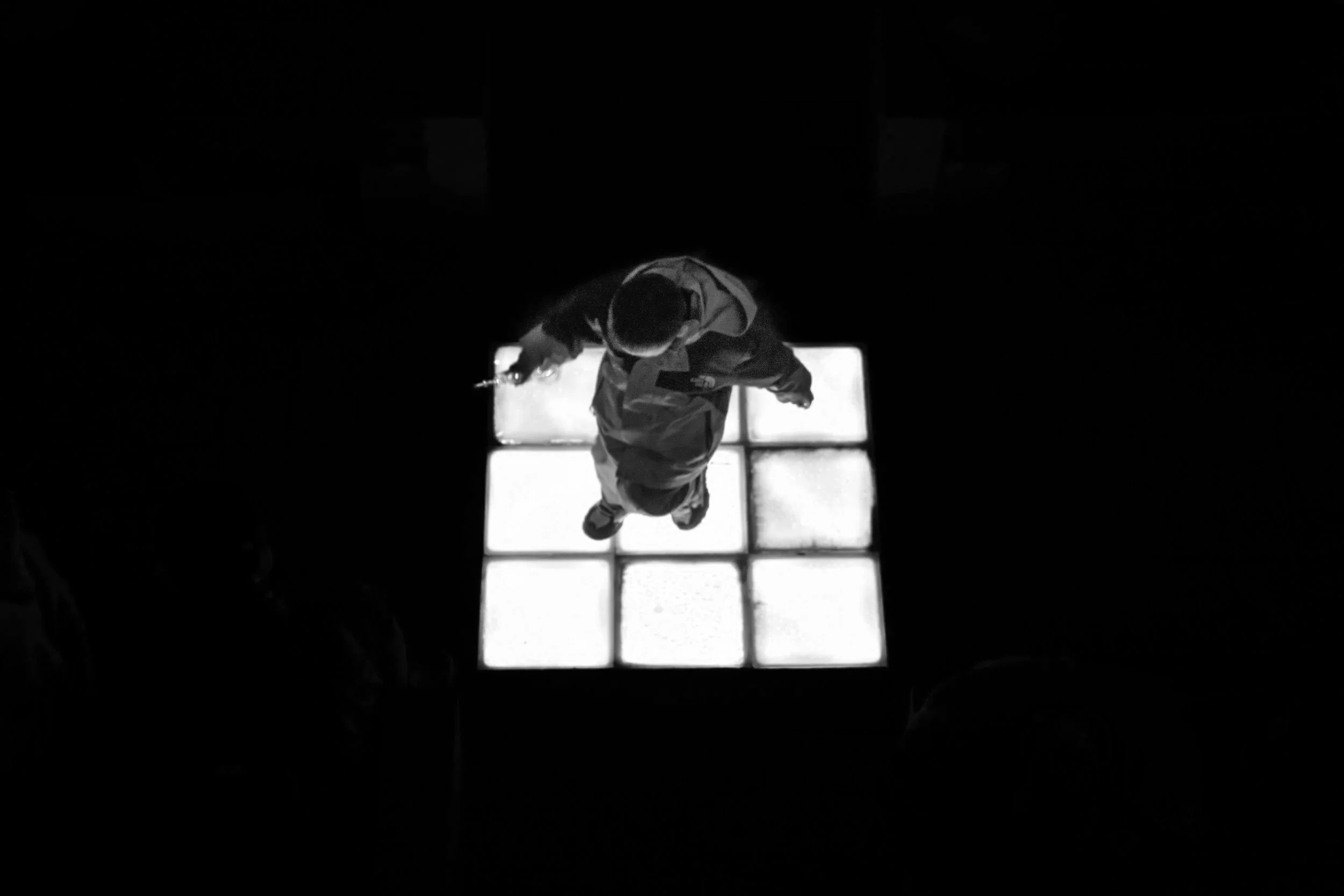 Overhead view of a person standing on illuminated yellow tiles in a dark space, holding a glowing object in one hand.