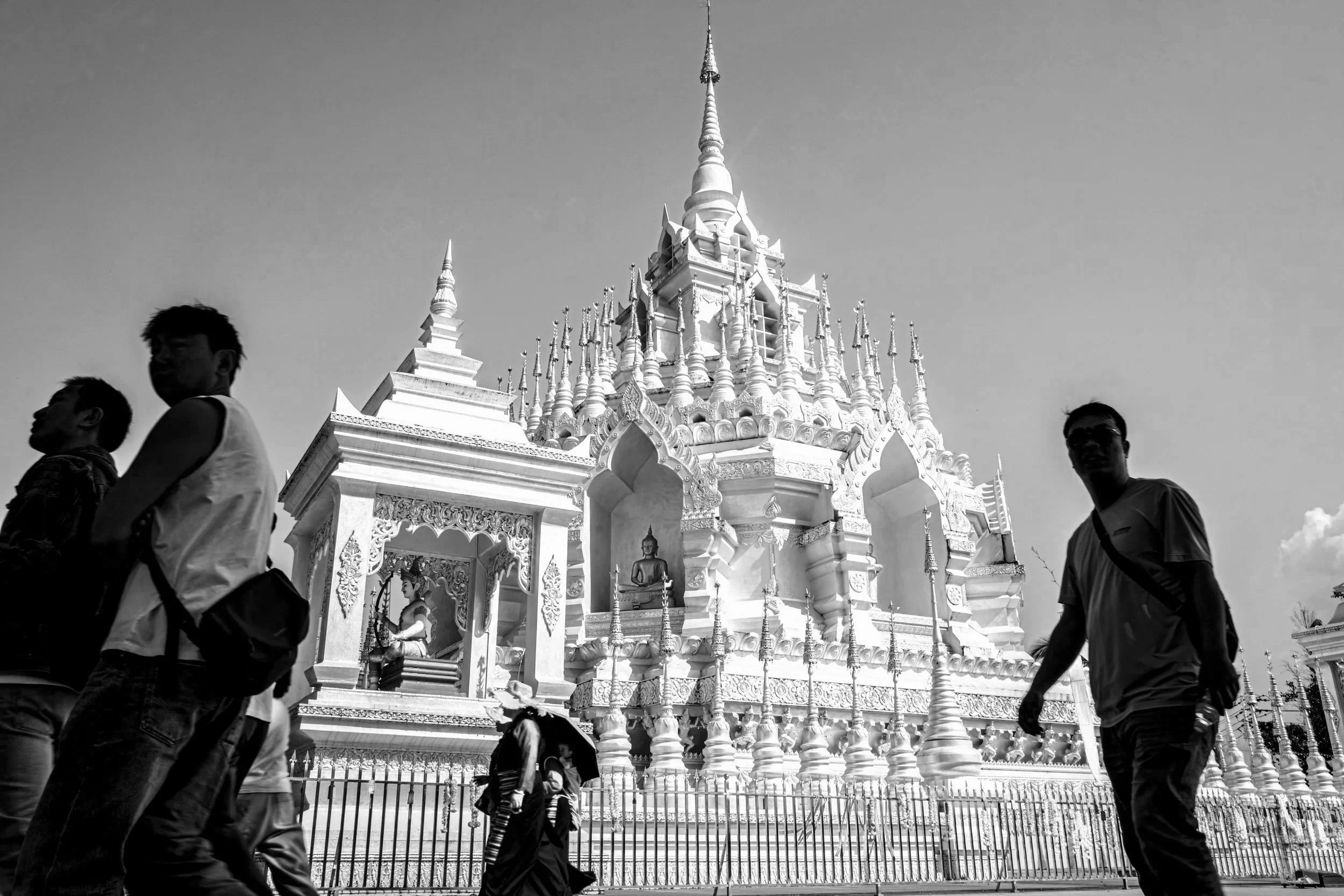 A white ornate temple with golden Buddha statues on its steps, surrounded by a black fence, and several people walking in front of it.