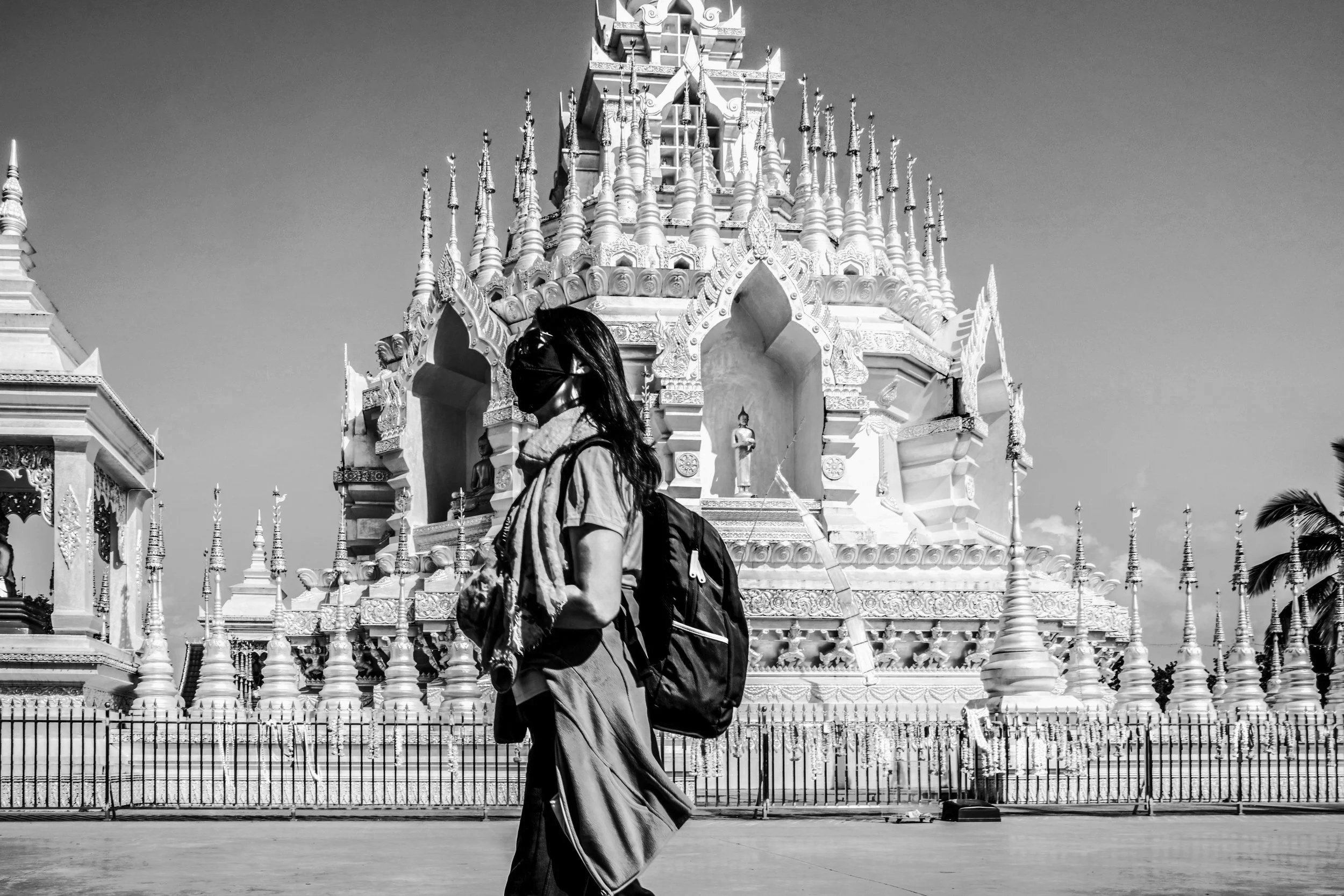 A person wearing a black mask, green shirt, and scarf around the neck, carrying a black backpack, walking past a large, ornate white temple with gold statues in the background.