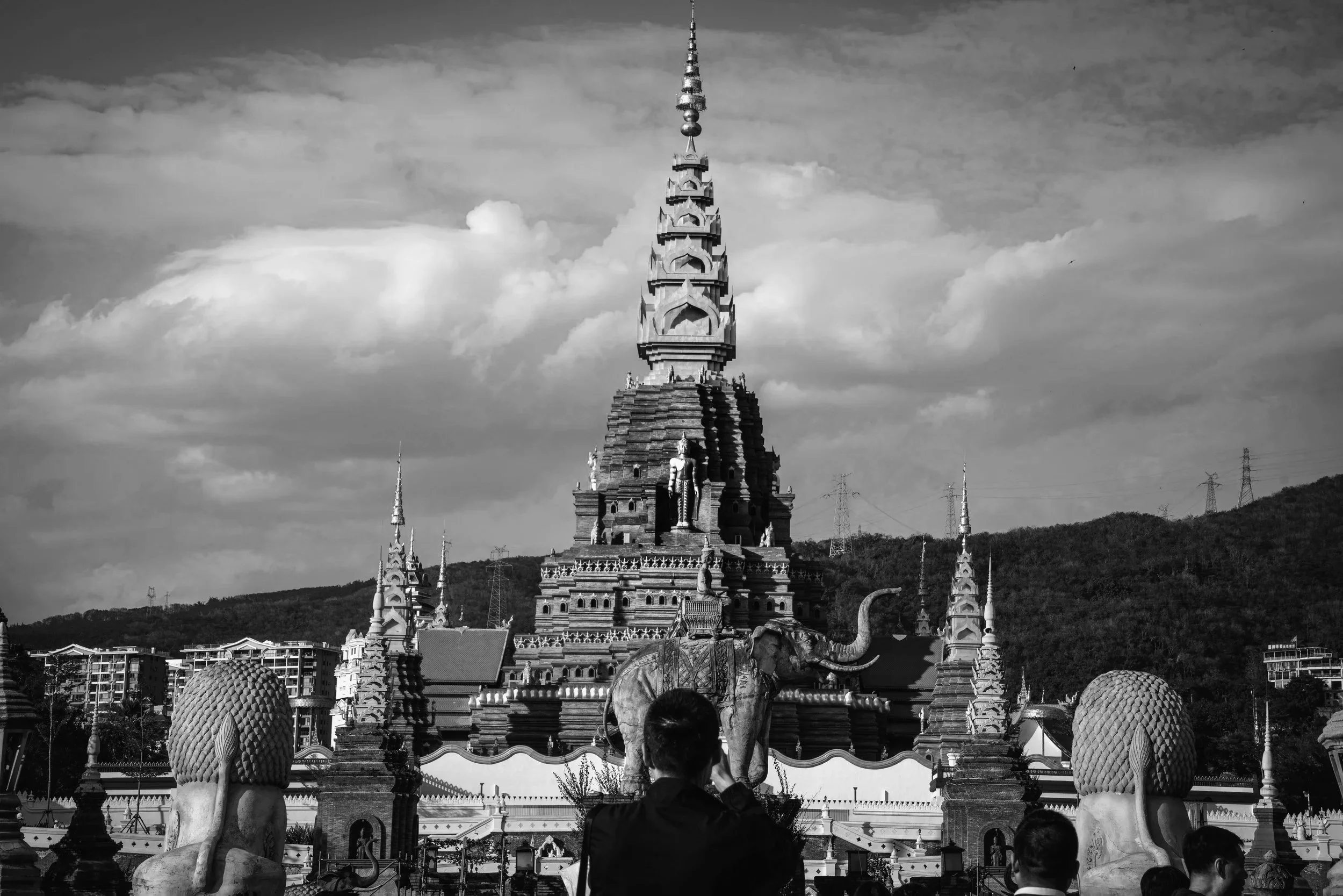 A person taking a photo of a Buddhist temple with a tall, multi-tiered pagoda, elephant statues, and ornate decoration, set against a cloudy sky and mountainous background.