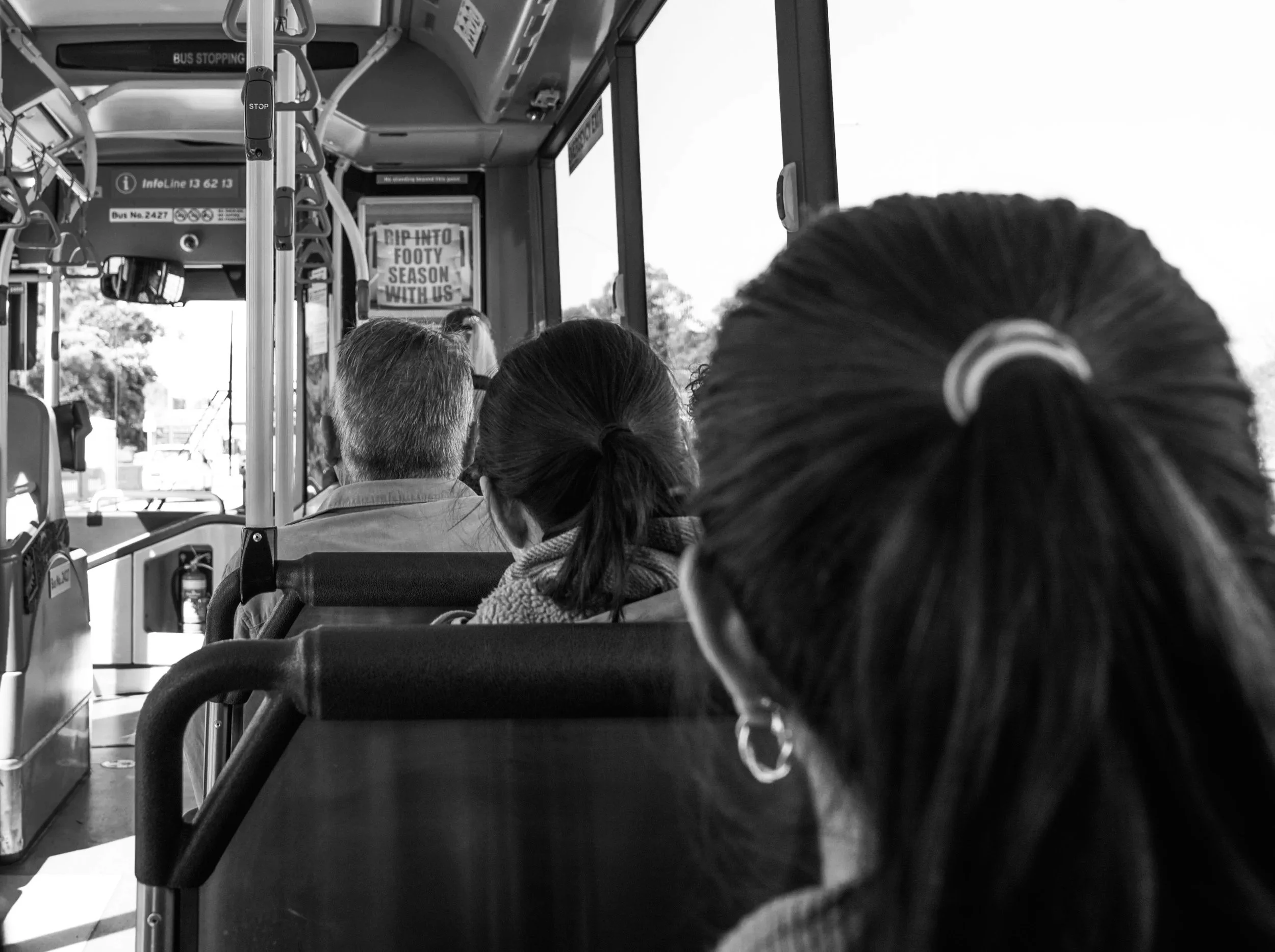 Black and white photo of people seated on a bus, viewed from the back, with a person in the foreground having dark hair in a ponytail. Others are seated facing forward, and a window on the right reveals trees and sky outside.