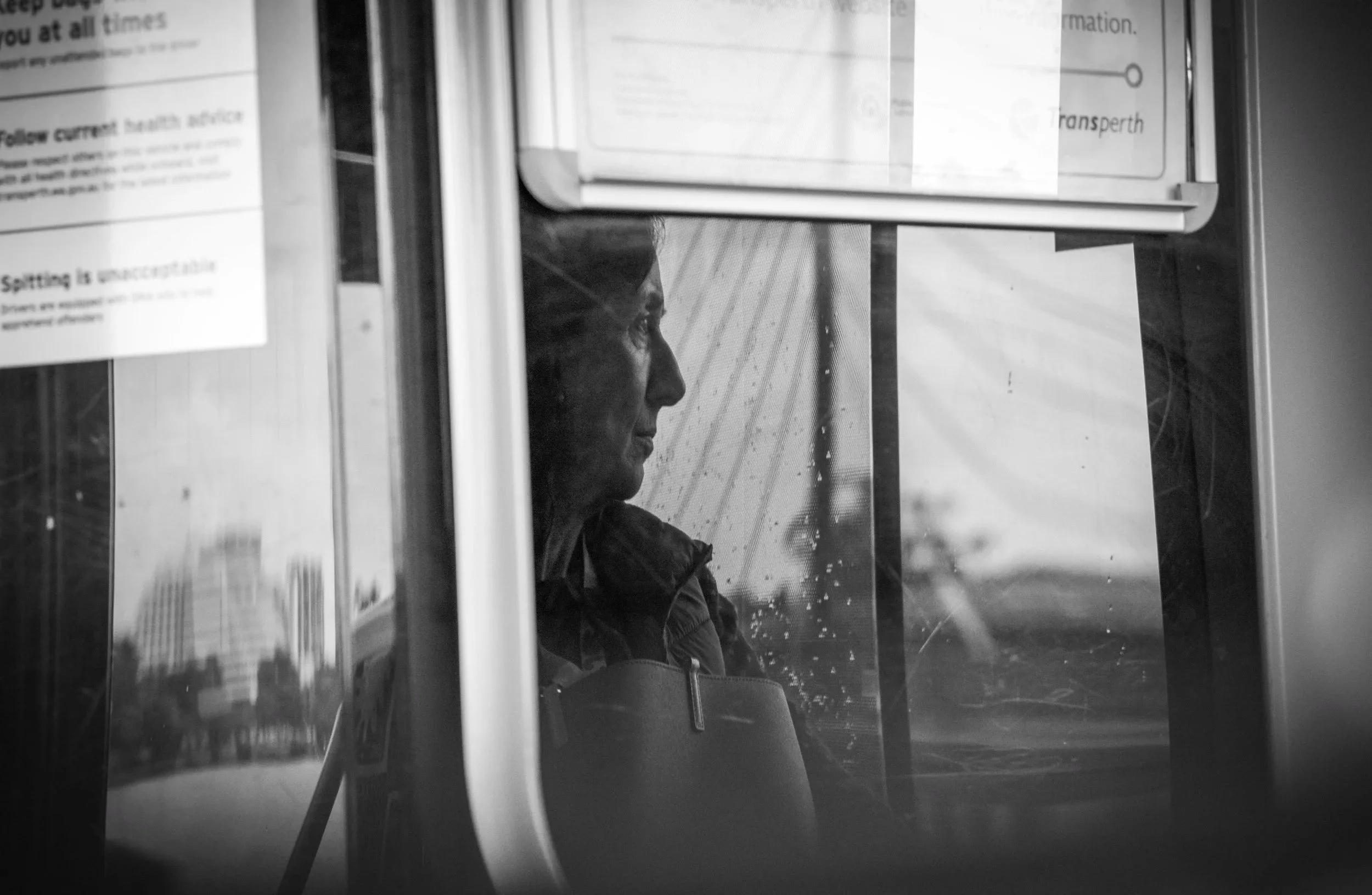 A person sitting inside a bus or train, seen through a window with raindrops, with city buildings visible in the background.