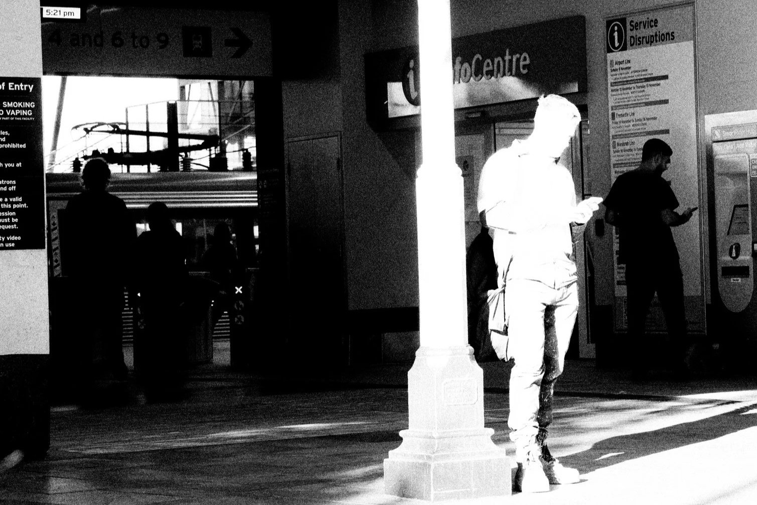 People walking and standing inside a busy train station with signs for tickets, info center, and platform information, in black and white.