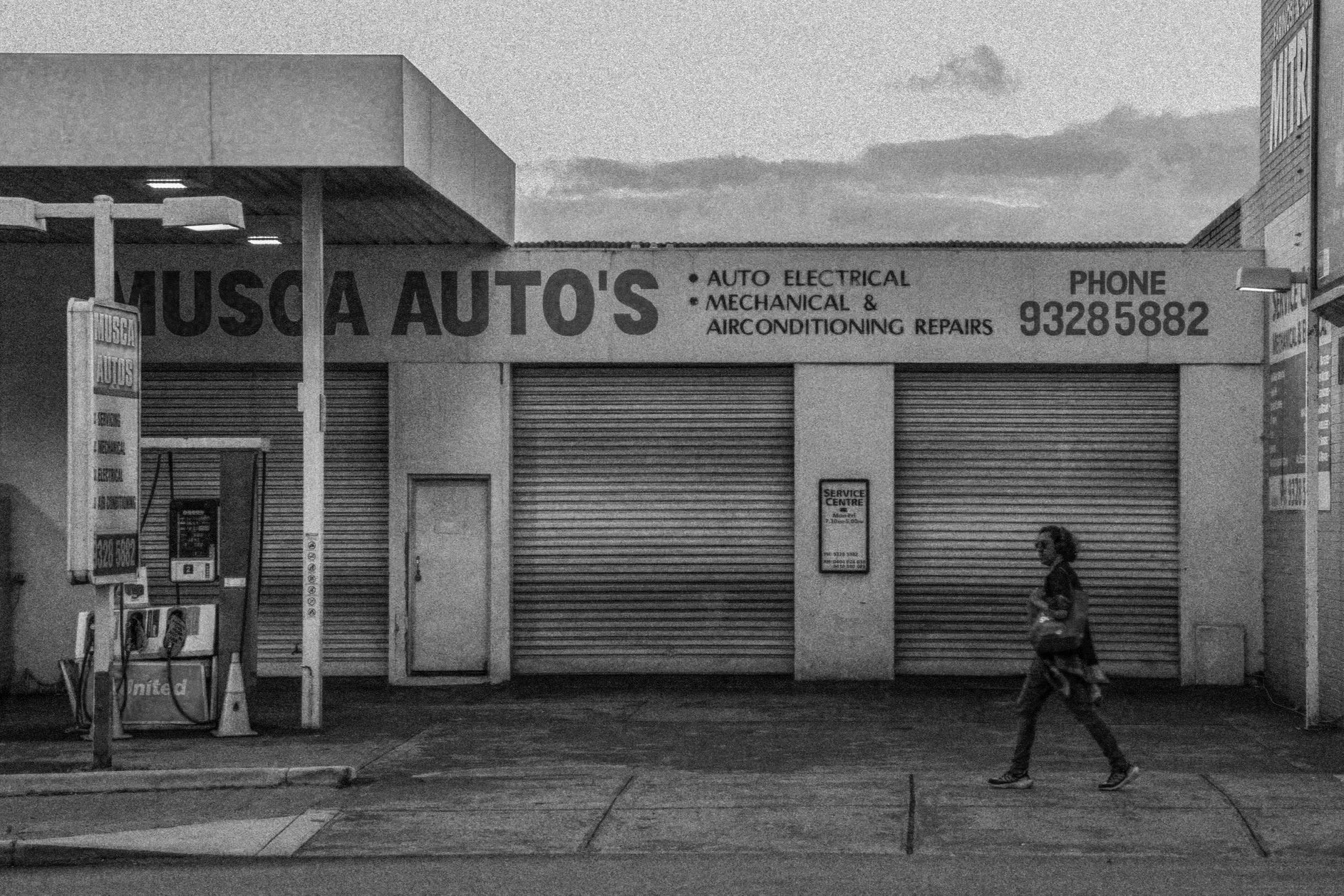 A building with a sign that reads 'Musca Auto's' and mentions auto electrical, mechanical, and air conditioning repairs, with a phone number '93285882'. There are closed roller shutters, a woman walking on the sidewalk, and a blue sky with some cloud