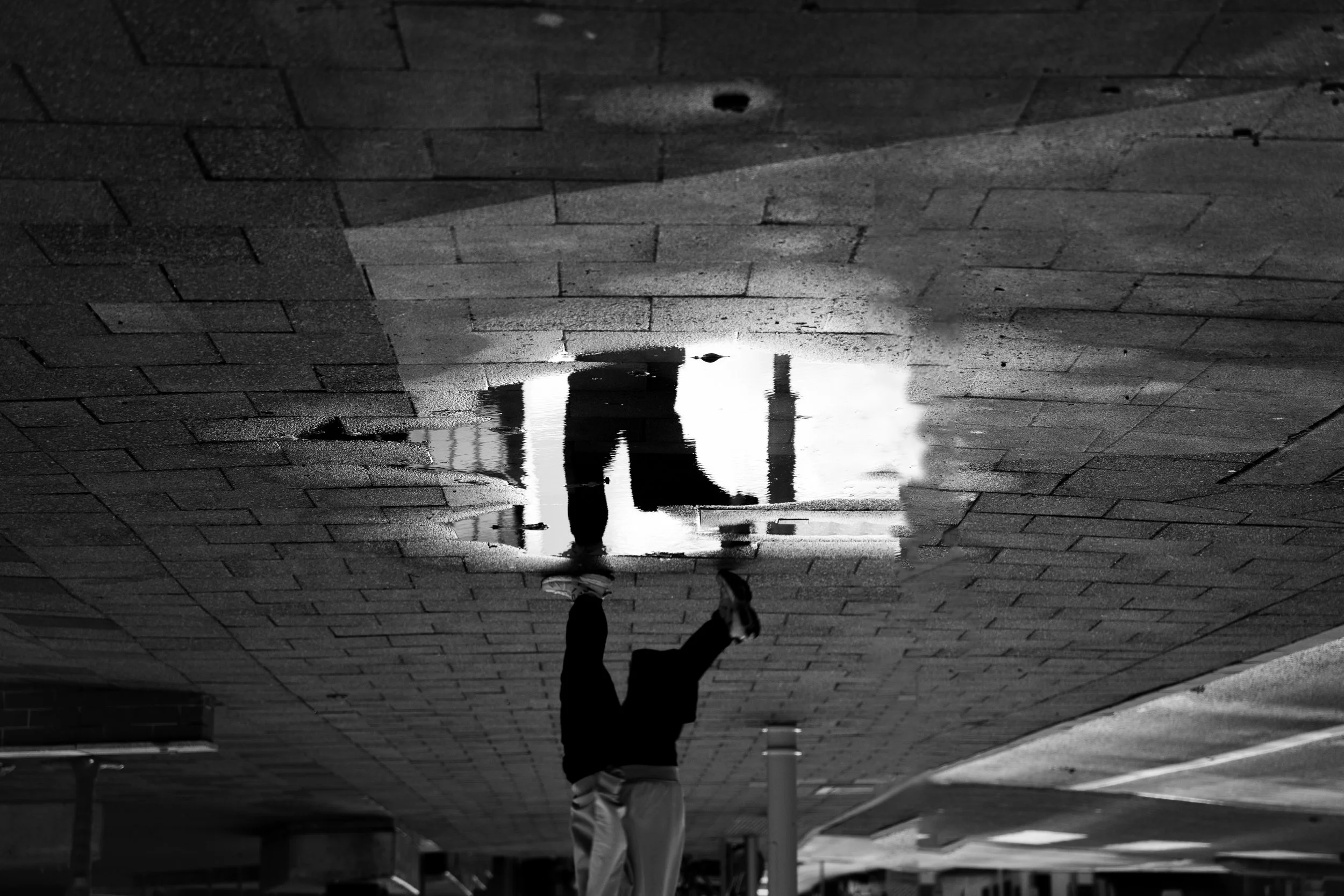 A person walking on a wet brick sidewalk, with their reflection visible in a puddle of water.