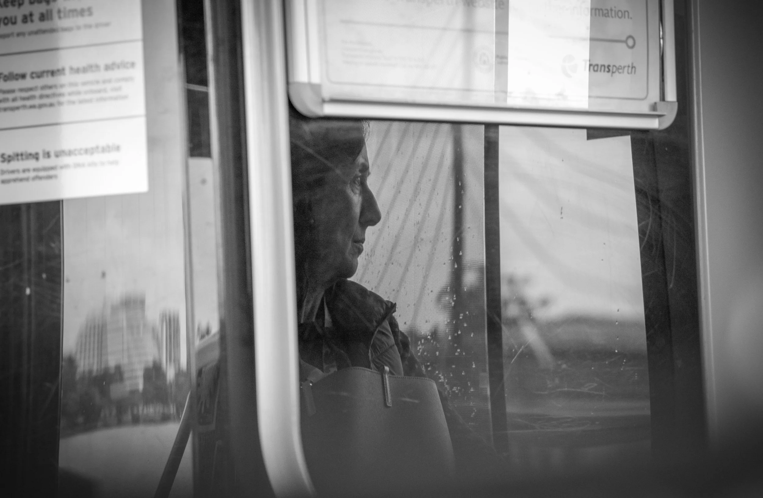 A person sitting inside a bus or train, seen through a window with raindrops, with city buildings visible in the background.